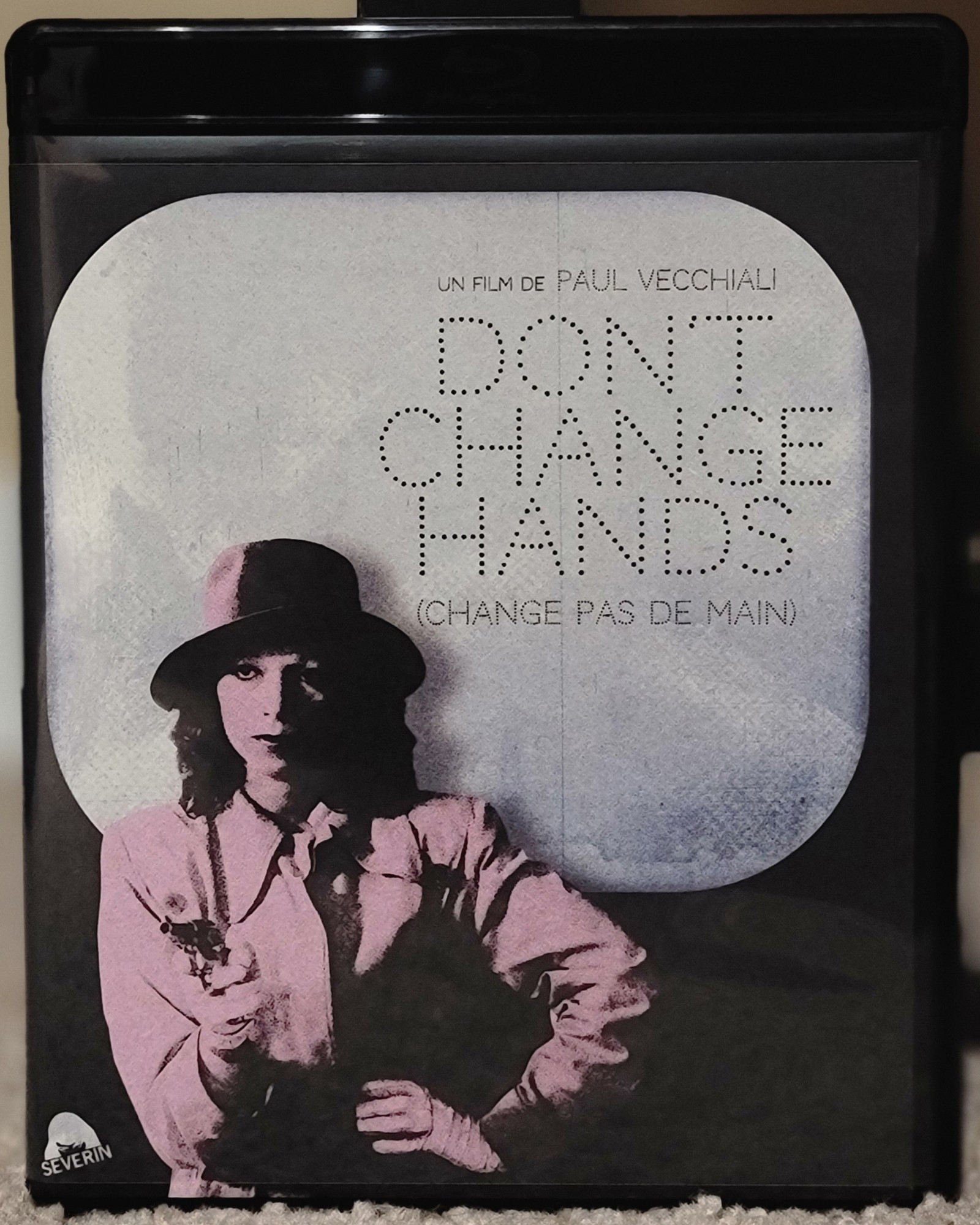 Blu ray cover for the film Don't Change Hands. The image art is of a woman, dresses as an old noir character would, holding a pistol with her other hand on her hip.