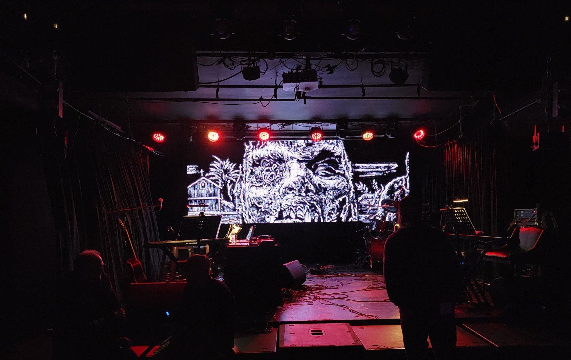 Concert stage with equipment and no people. A big screen has a pixilated image of a zombie.