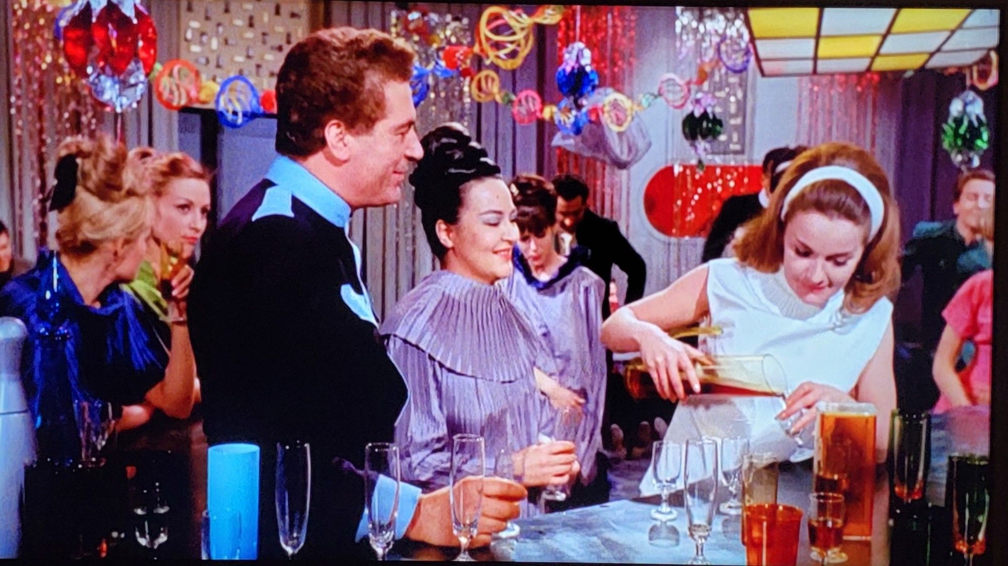 People celebrating at a party. They're wearing a 1960s idea of futuristic clothes. The room is filled with colorful party streamers and decor.