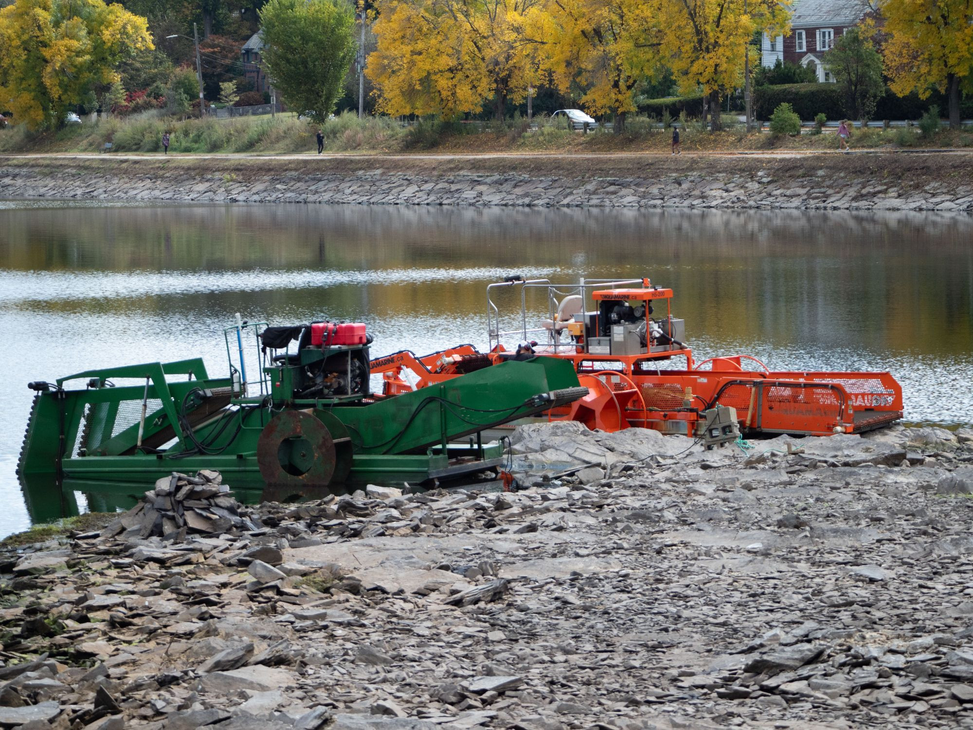 Two dredging machines, one green and one red, moored on a reservoir's rocky shore. In the background the path wrapping around the reservoir is visible, as are some autumnal trees.