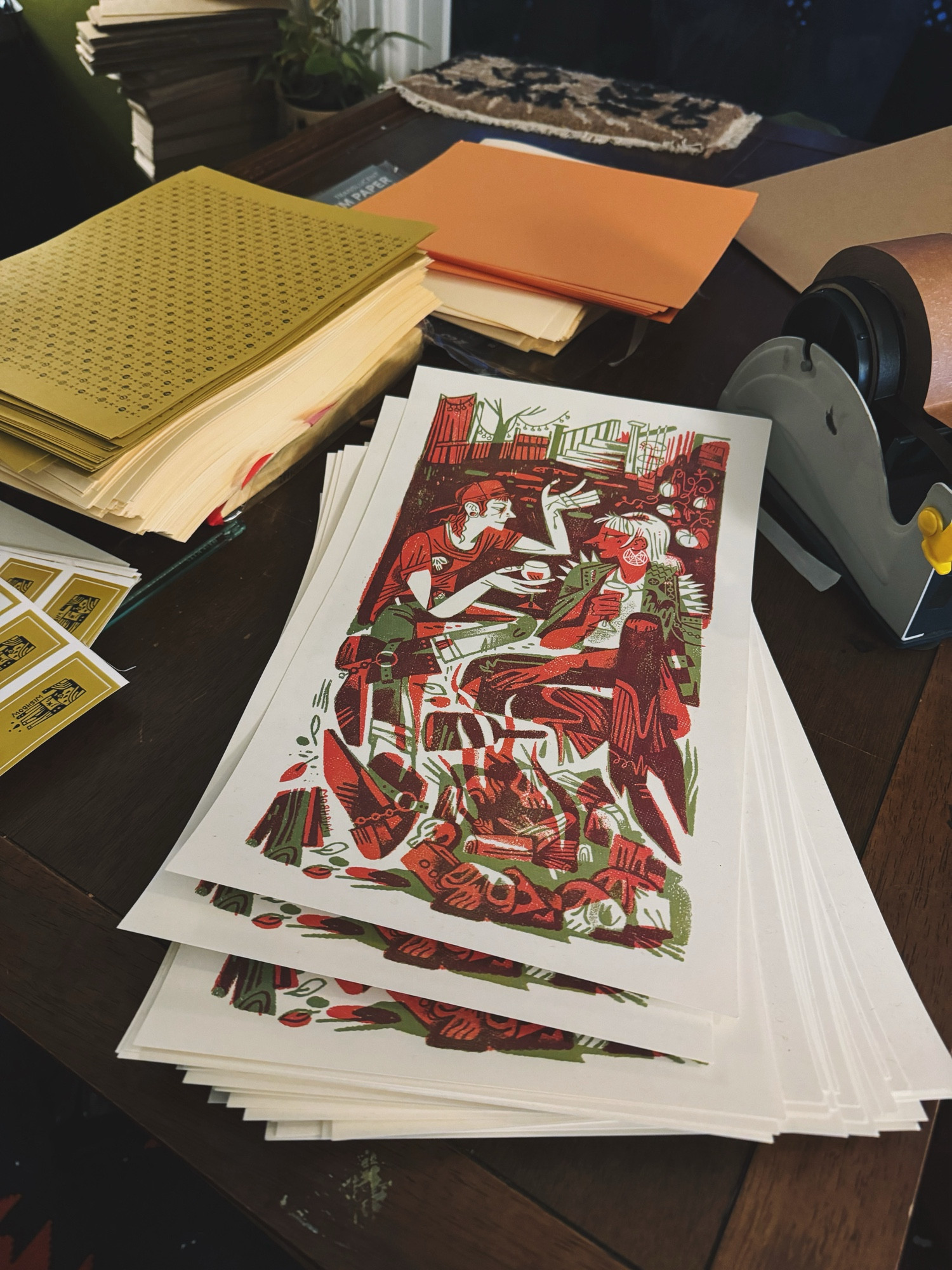 Piles of paper on a desk including a print of two people around a fire drinking wine