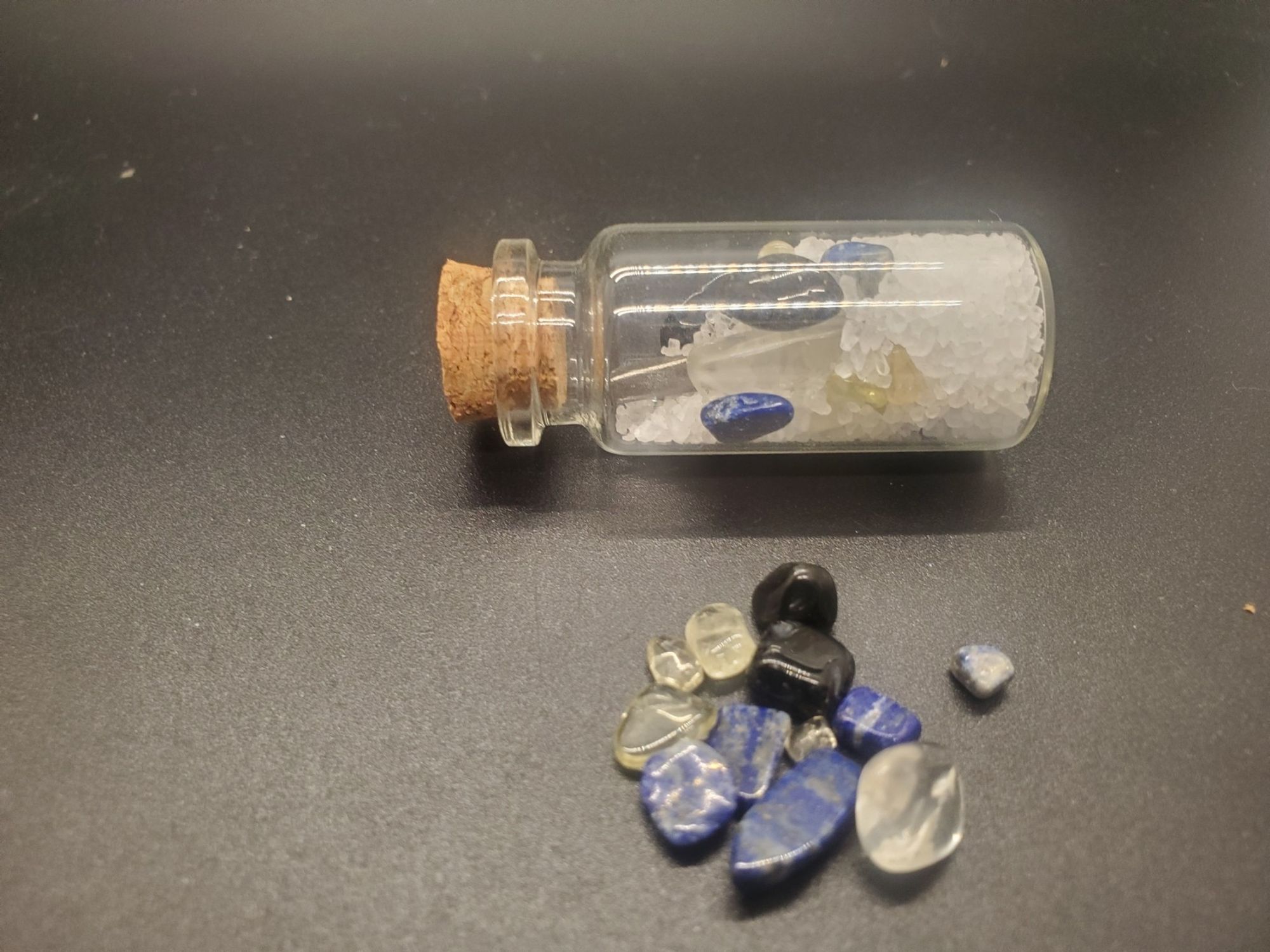 a small jar lies beside a pile of crystals. In side the jar those crystals (Citrine, Lapis Lazuli, and Obsidian) are mixed with Salt and Clear Quartz with an Iron nail through the middle of the mixture.