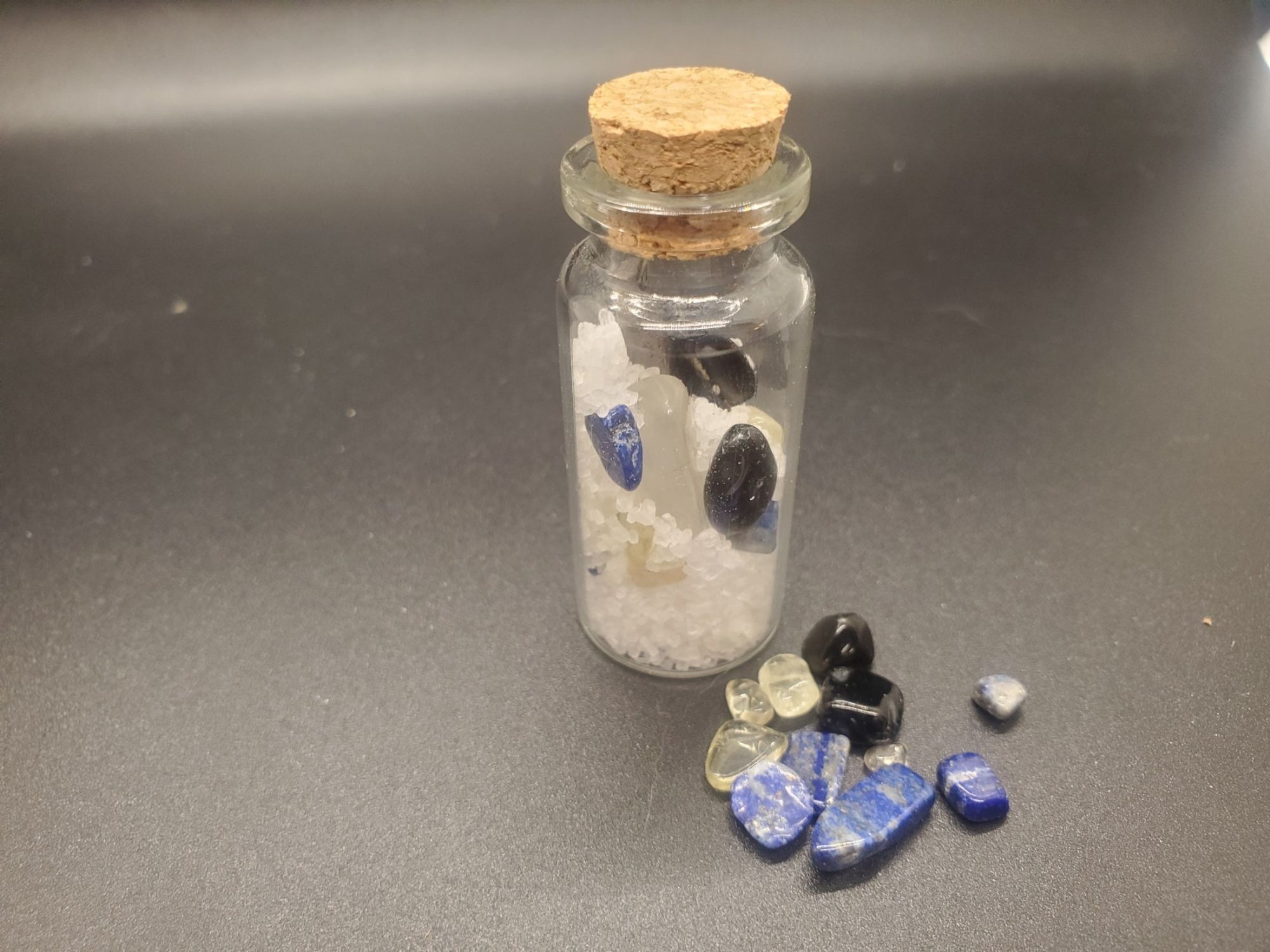 a small jar sits beside a pile of crystals. In side the jar those crystals (Citrine, Lapis Lazuli, and Obsidian) are mixed with Salt and Clear Quartz with an Iron nail through the middle of the mixture.