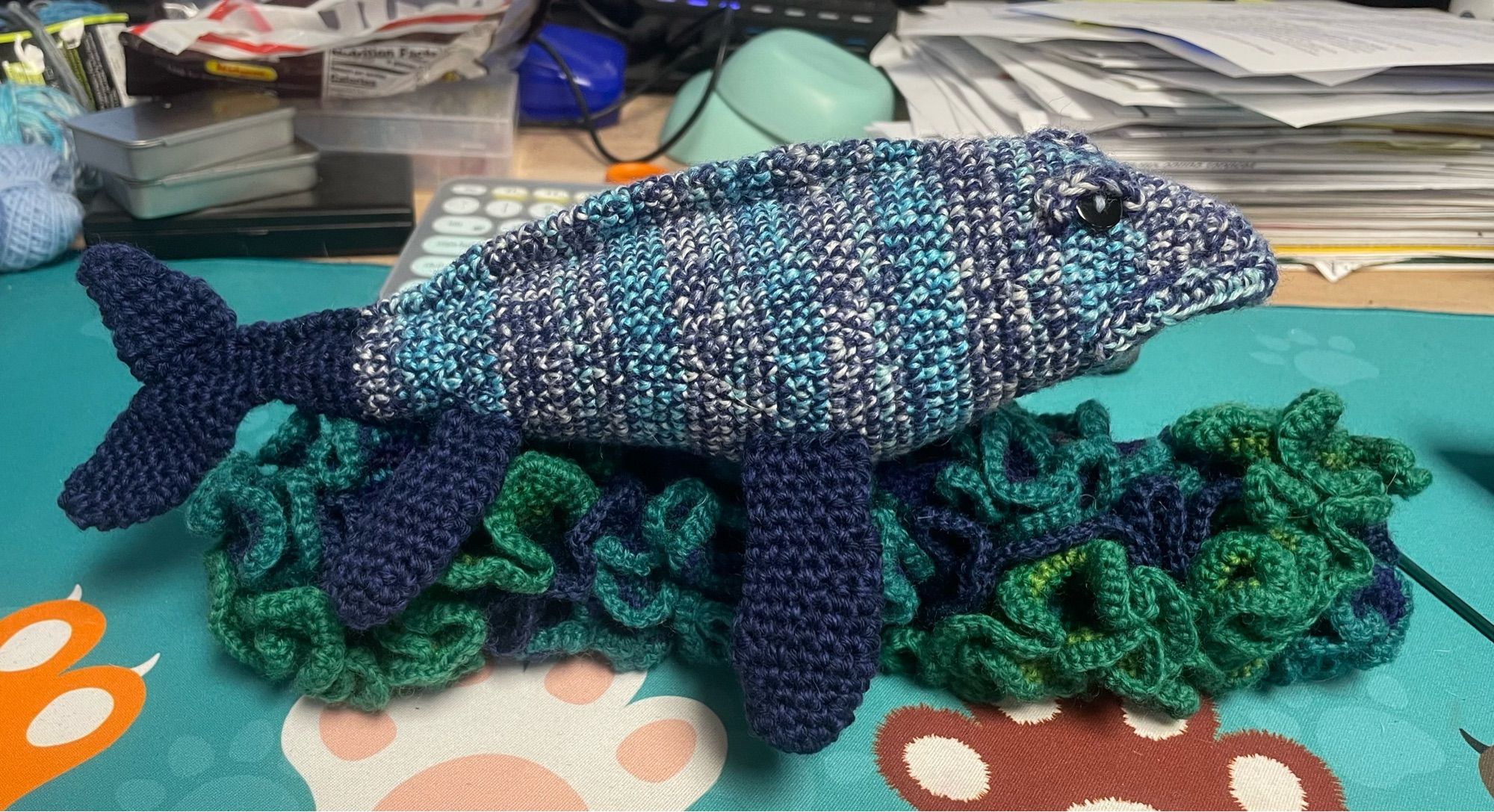 A blue and blue and white crocheted Mosasaur with navy blue fins and tail supported on a blue, teal, and green crocheted coral. The mosasaur‘s body is stripes of marled navy & white and navy & light blue. Its eyes are black buttons.