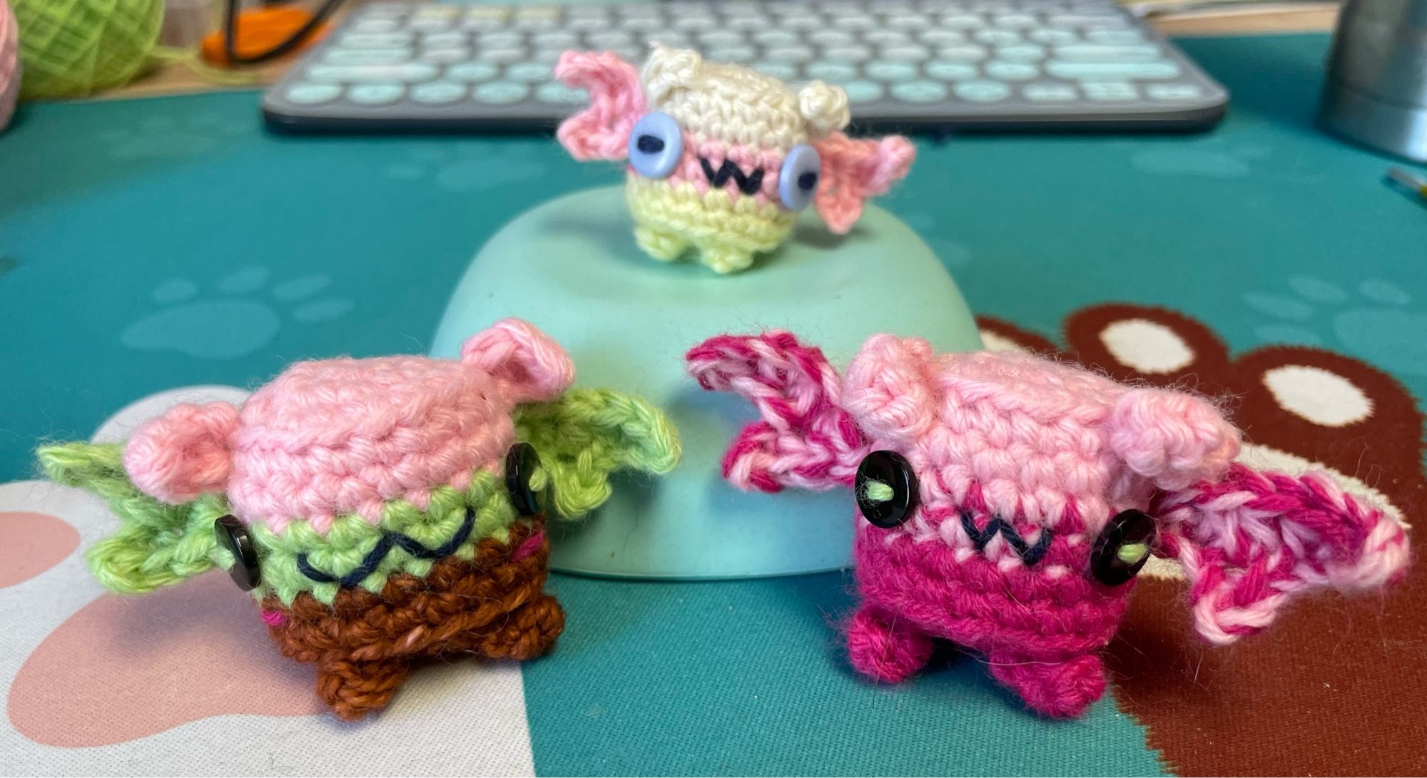 Tiny crocheter Marshmallow bat on top of a thing, with larger friends Spumoni bat & Raspberry Sorbet bat. Spumoni Ice Cream is pink on top, green in the middle, and chocolate brown on the bottom. Raspberry Sorbet is pink  on top, dark pink on the bottom, and the two shades marled together in the middle. They both have black button eyes. Their ears, wings, and feet match, respectively, their top, middle, and bottom colors. They al have W mouths.