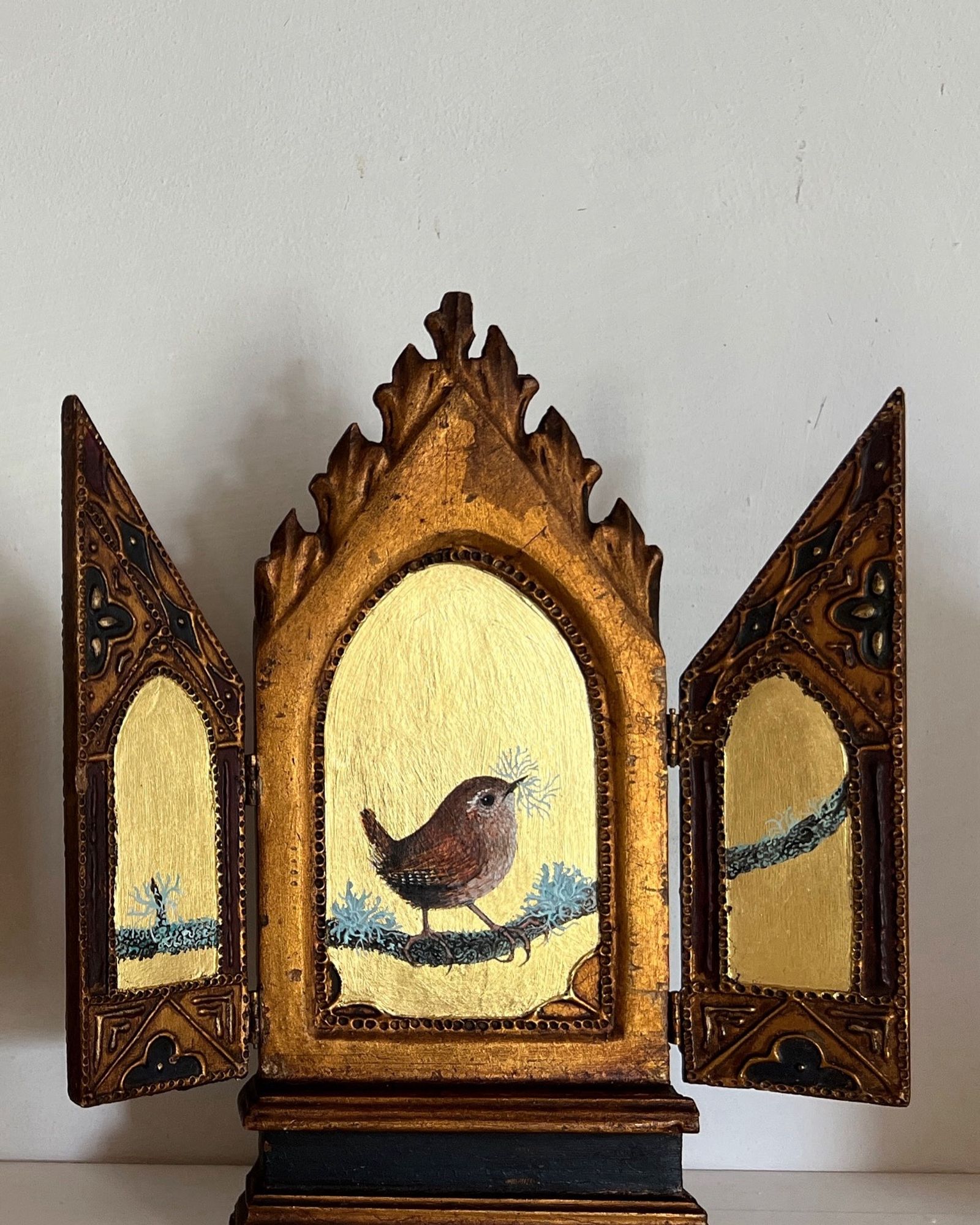 A painted triptych of a wren perched on a twig with lichens. The background of the painting is gold leaf, which is laid on antique French carved wood and gesso folding panels. The wren, which is facing to the right has a little bundle of lichens in his bill, for nest building