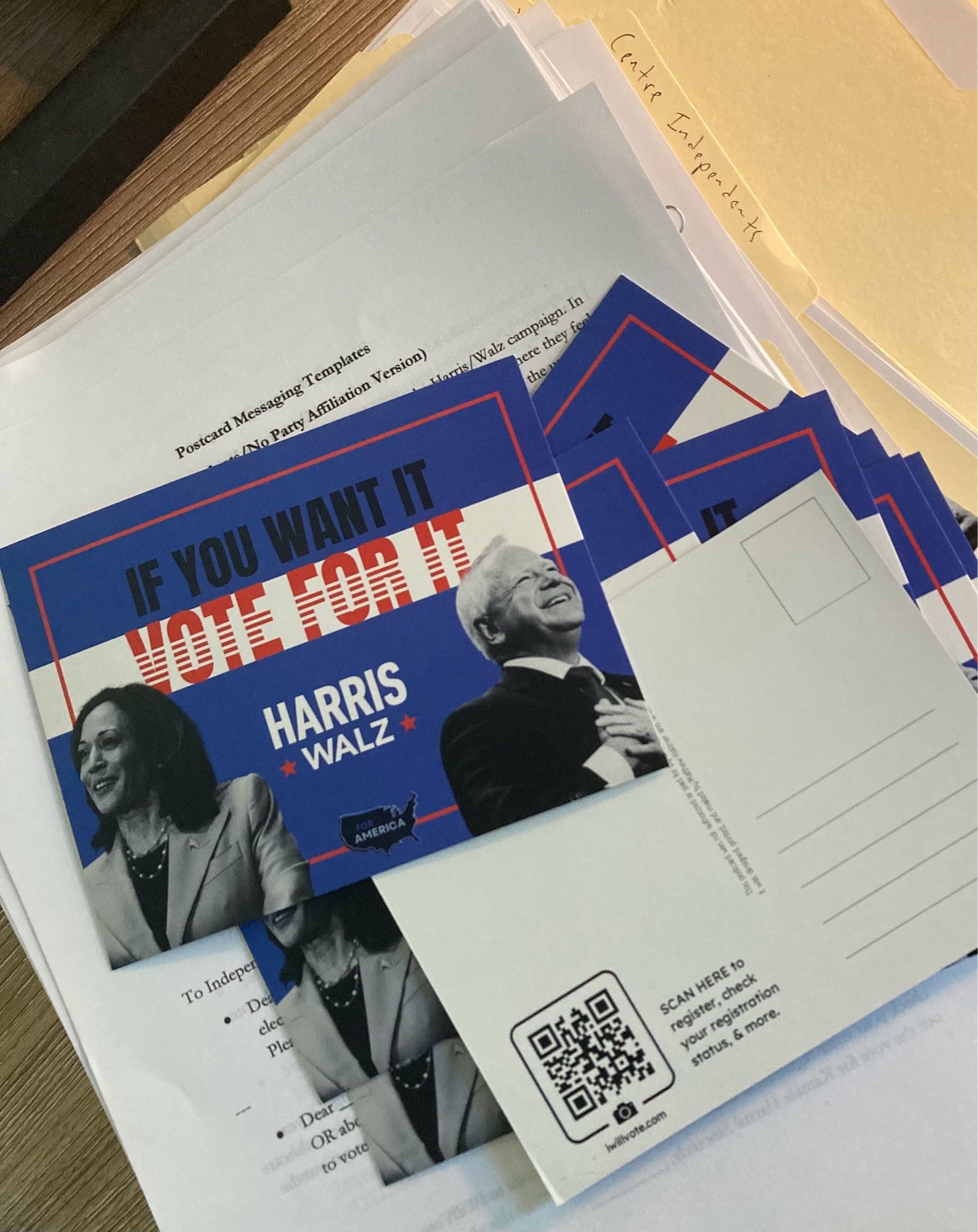 A pile of postcards strewn over paper and folders. The postcards read: “If you want it, vote for it” and “Harris Walz”. One postcard is flipped over, showing the backside with a QR code to Iwillvote.com and space to write messages to potential voters.