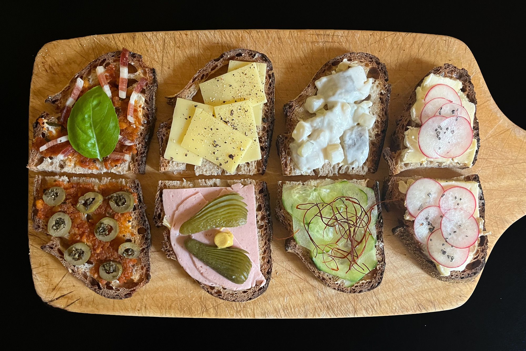 Auf einem Holzbrett acht einander paarweise zugeordnete Brotscheiben, Belag: 
– Butter, rotes Pesto, Speck, Basilikum / Kräuteroliven 
– Butter, Gouda,  Pfeffer / Lyoner, Gewürzgurke, Senf
– Butter, Heringssalat / Salatgurke, Chilifäden
– Butter, Kuhweichkäse, Radischen, Vulkansalz