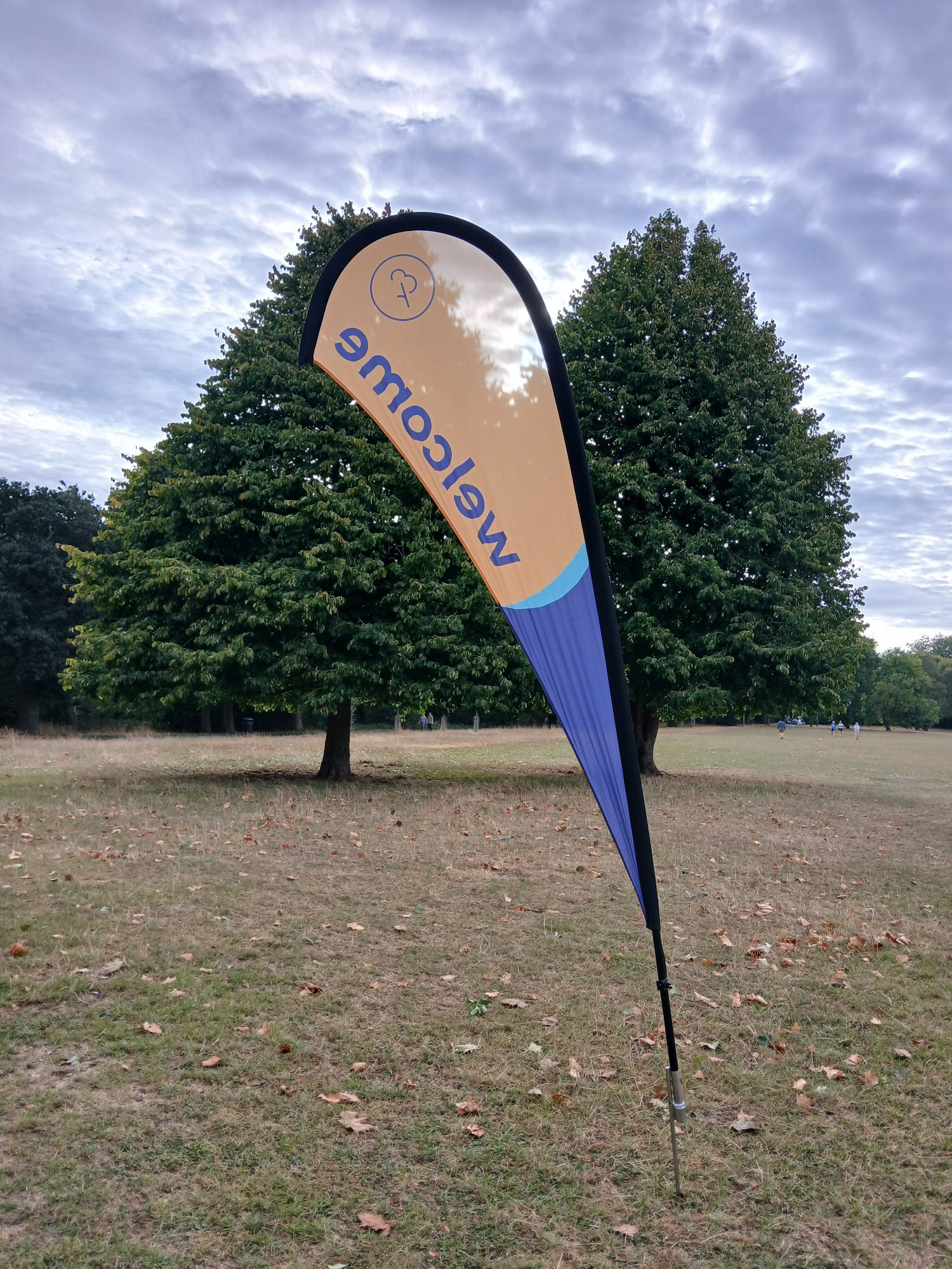 Photo of a ParkRun welcome banner in front of two trees in a parched park. The trees are still in full, green leaf but a scattering of brown leaves are lying on the ground, blending in with bare earth and dry grass.