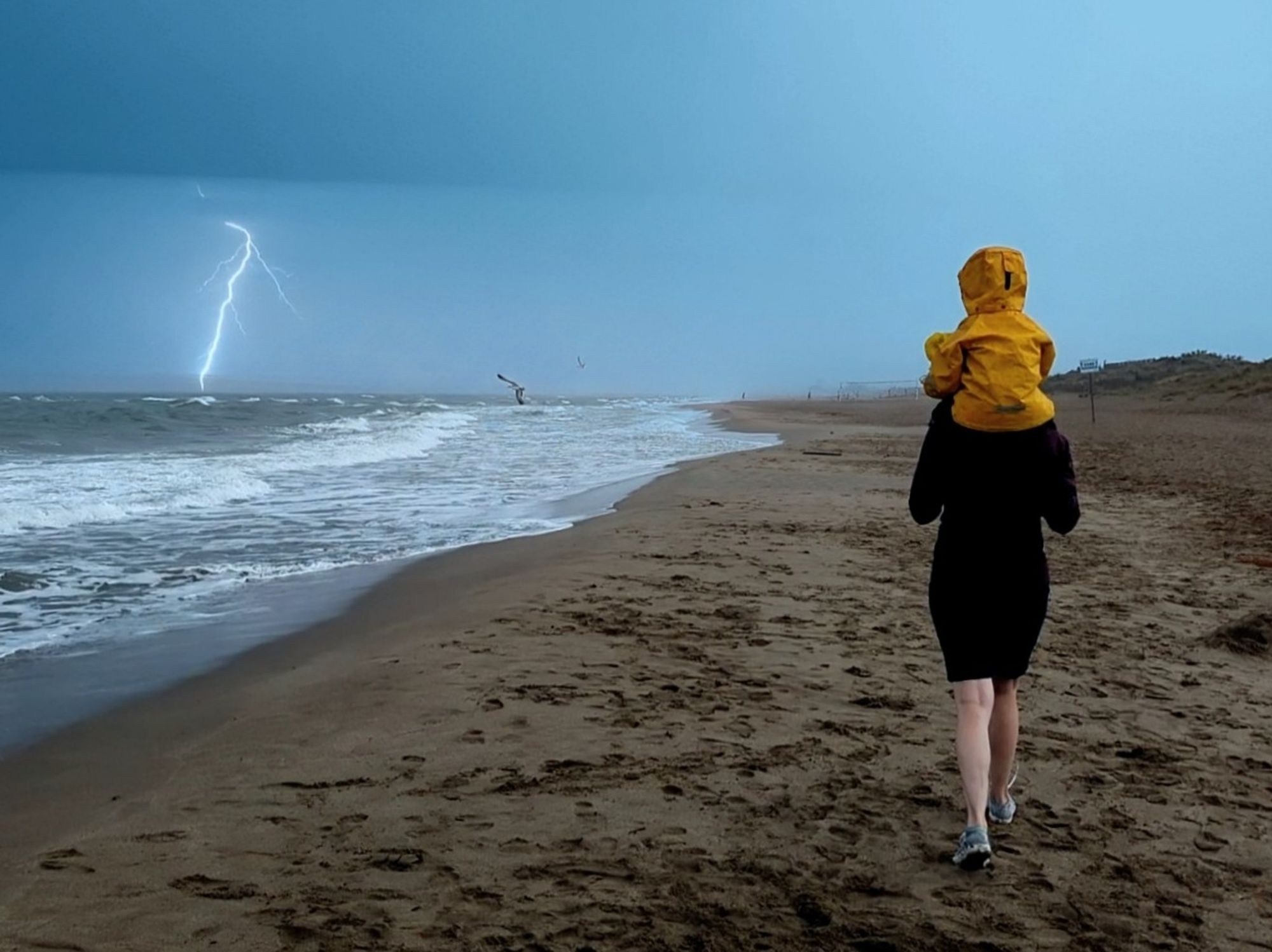 Picture of a woman walking on a rainy beach with a child on their shoulders. There is a bolt of lightning in the background