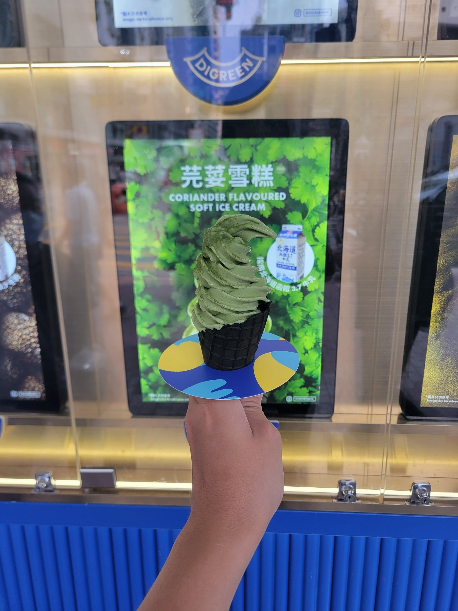 A coriander flavoured, green soft-serve ice cream served in a dark green waffle cone, with a promotional poster promoting said ice cream in the background.