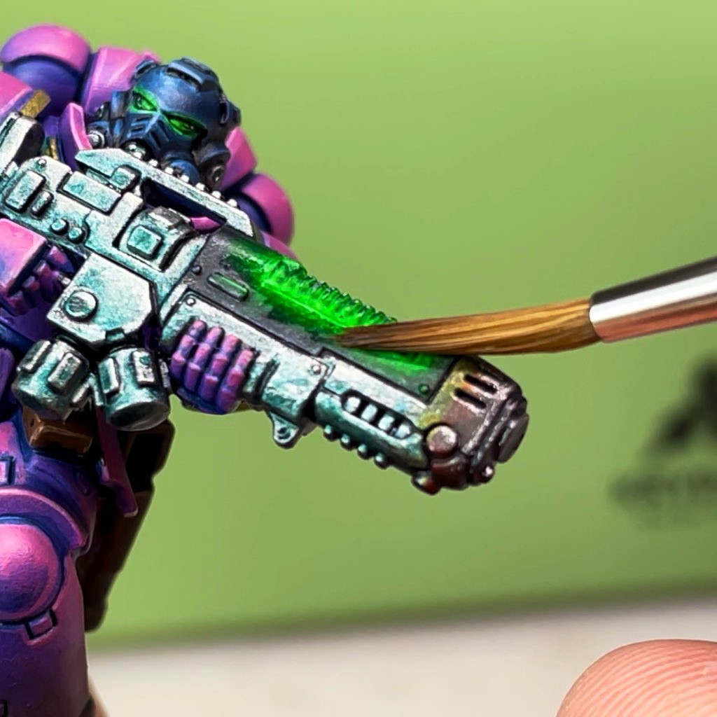 “Close-up of a Warhammer 40k Space Marine miniature being painted. The model features vibrant pink and purple power armor with metallic details. The image highlights a glowing green plasma effect being applied to the weapon’s core using a fine brush. The intricate shading and highlighting techniques emphasize a futuristic, cyberpunk aesthetic. Ideal for hobbyists and miniature painters seeking inspiration for Warhammer 40k painting techniques and plasma glow effects.”