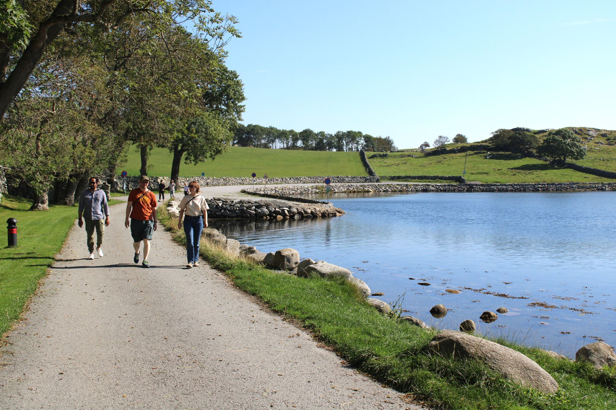 Three people walking up a path next to a bay surrounded by green fields.