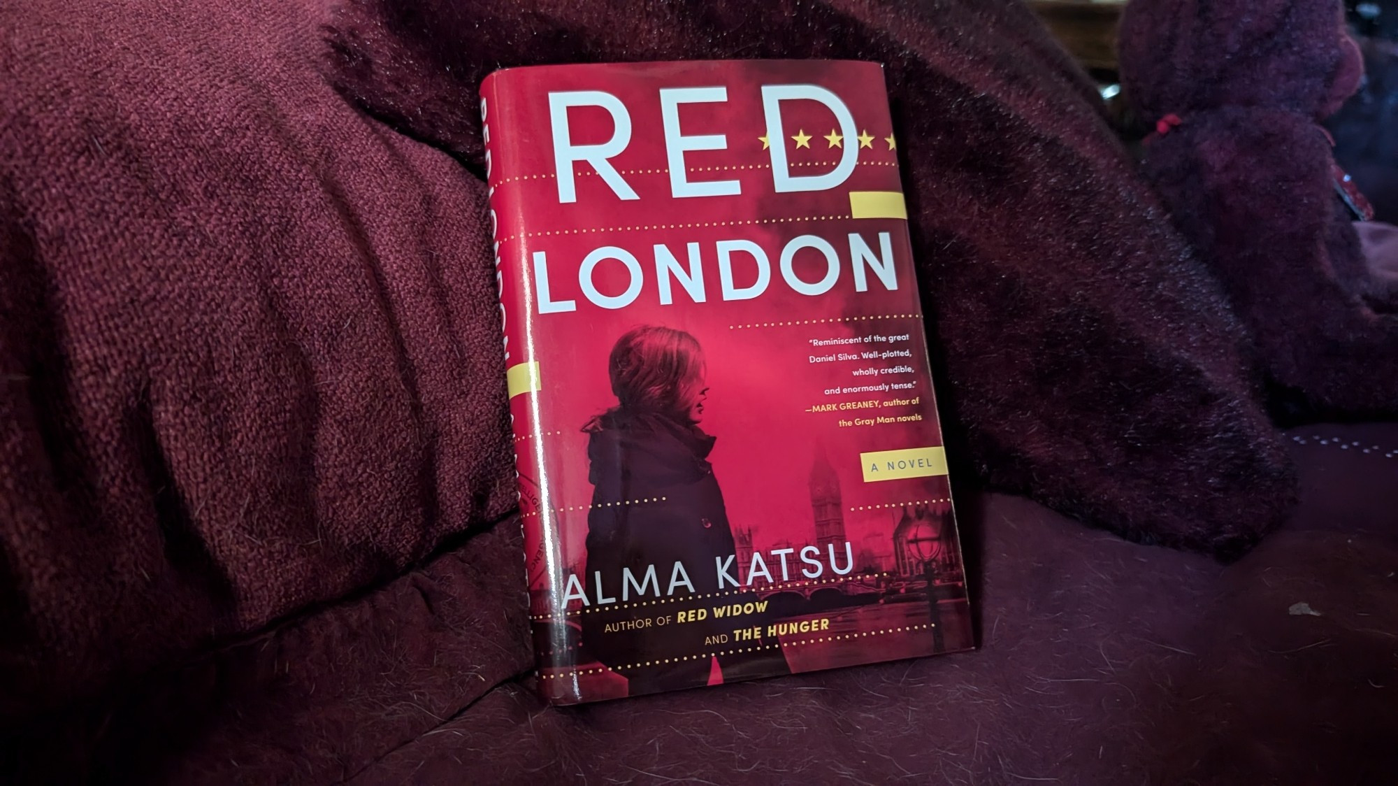 The cover is red, featuring the profile of a woman in a coat before a London skyline across a river.  They're depicted in black, like monochrome, but with red instead of white. A few small yellow rectangles poke in from the edges, and five little yellow stars sit in a line near the top right.