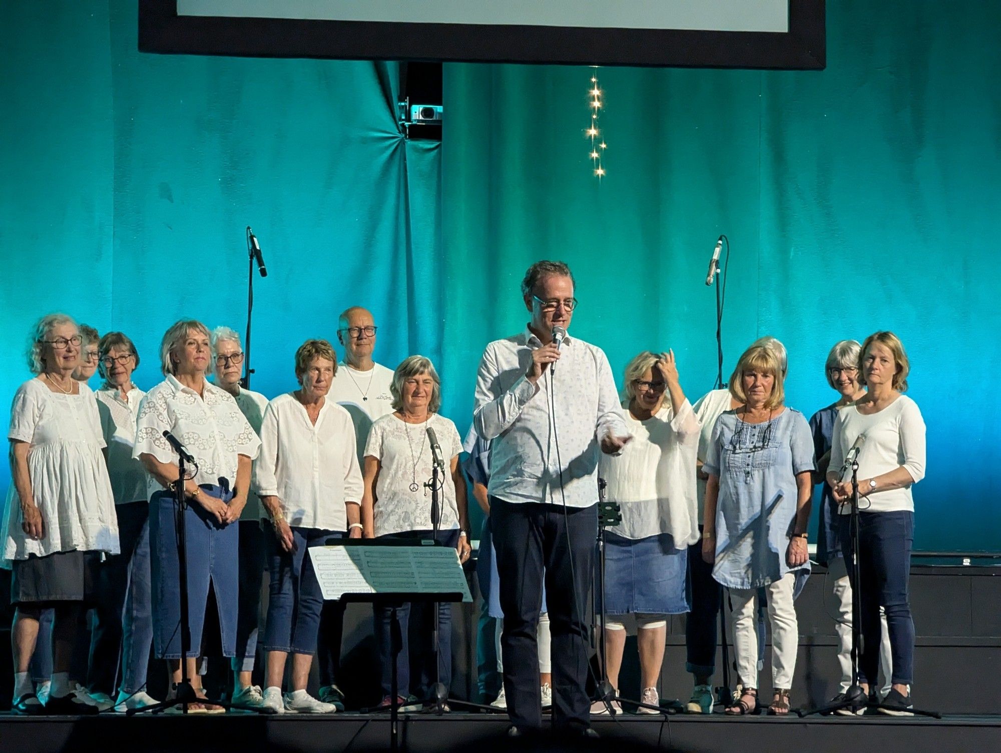 Photograph of Gospel Journey Choir performance at Jubilee Central in Hull. The choir are wearing white tops and blue jeans or skirts. They are standing in front of a glue-green curtain backdrop and there are white fairy lights hanging down in the centre of the picture. The director, Joakim Arenius, is speaking to the audience who are sitting out of shot.