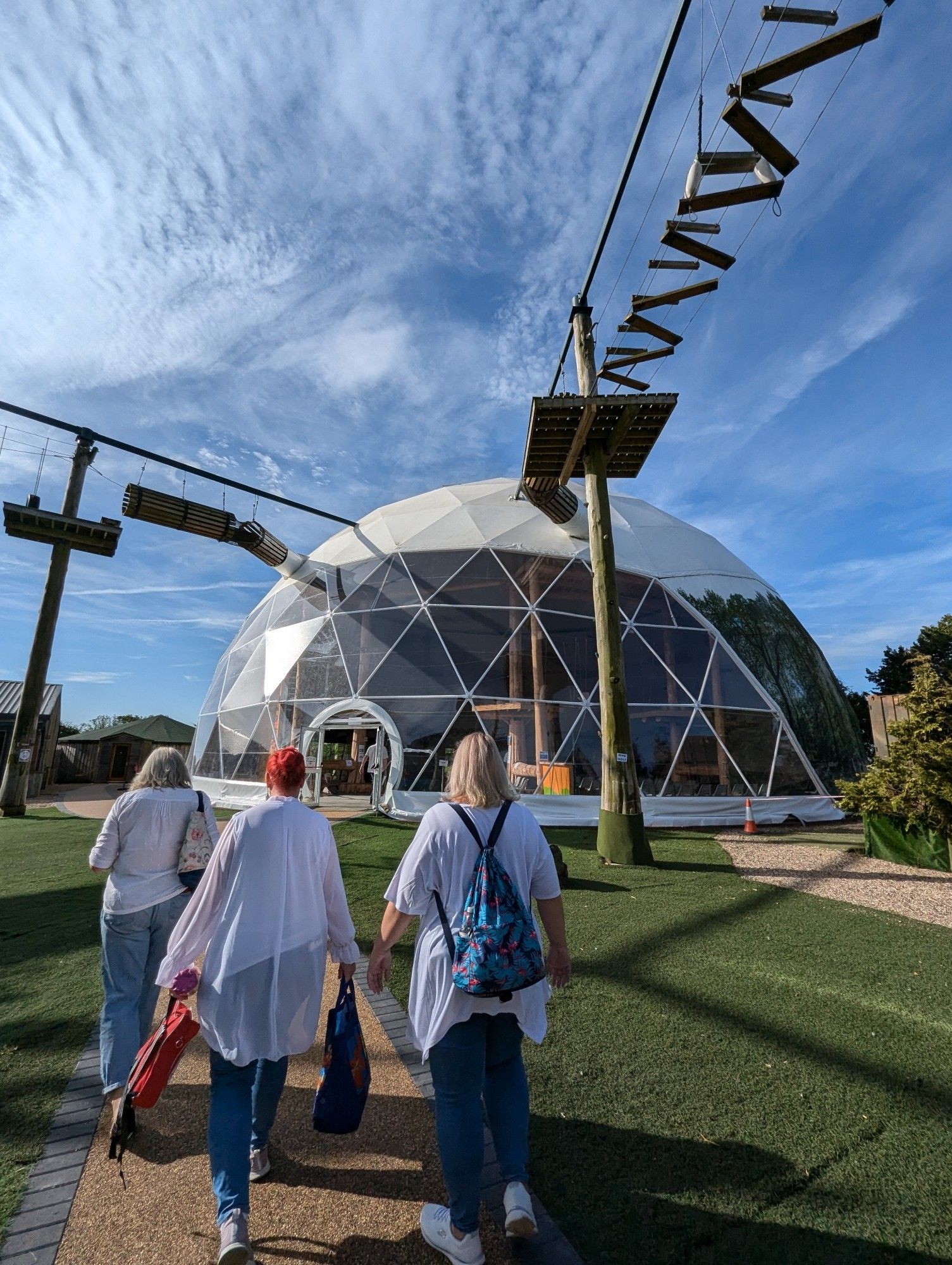 Photograph of Let Loose Adventure Park Geodome, where Revive Church hold worship, with FTLOG founder Helen Garnett and members of AFO Choir and Hotgospel dresses in blue jeans and white tops walking away from the camera towards the Geodome. The day is sunny and the sky is blue. There are high-level walkways around the site.