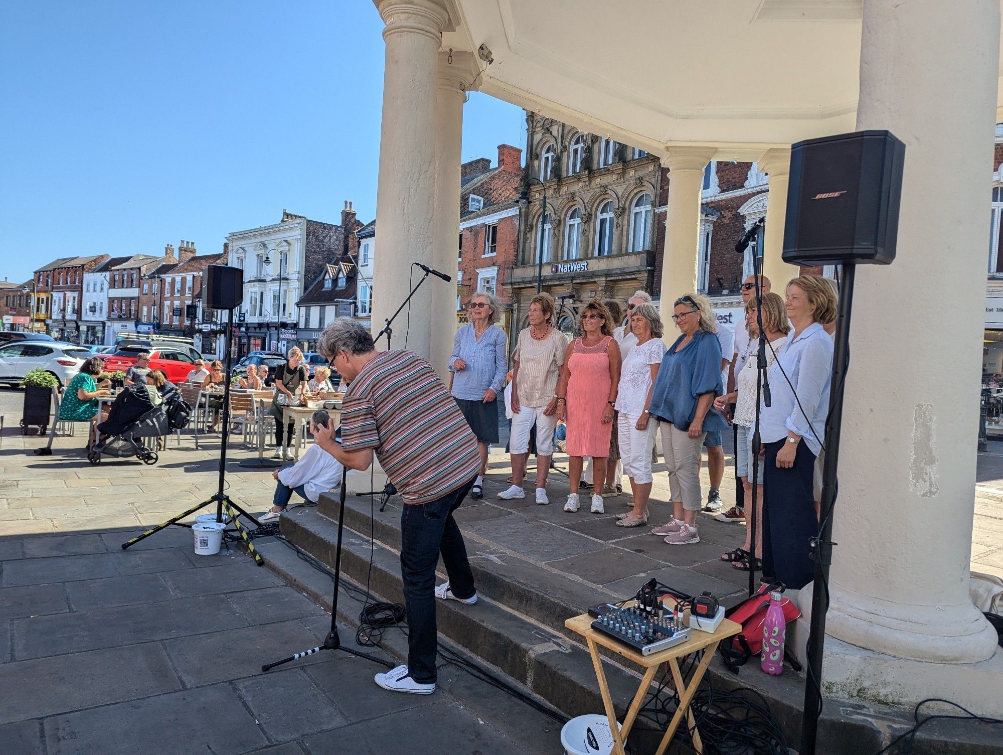 Photograph of Gospel Journey Choir busking in Beverley. They are standing under the canopy at Market Cross. There is audio equipment set up around the choir - speakers, mics and a small mixing desk. People are sitting at cafe tables in the background. The mid afternoon sunshine casts shadows and the sky is blue. There are buildings - shops and pubs - further in the background.