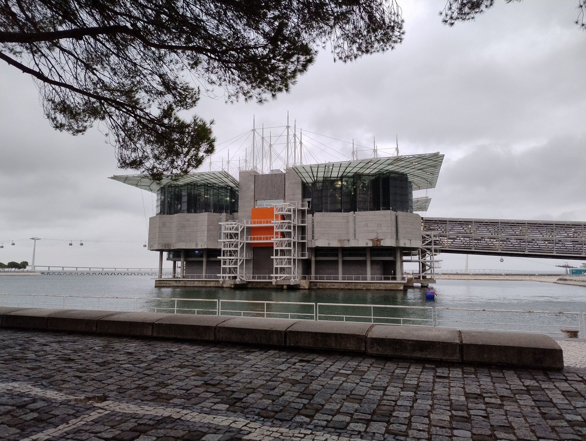 ein grosses gebäude mit quadratischem grundriss, das eine künstliche insel darstellt. es hat drei etagen (jede ca 3 mal die höhe einer moderen wohnung) die unterste hat ein nach innen versetztes, betonquadrat. aussen rum gehen betonsäulen. es erinnert an eine ölplattform. die zweite etage besteht aus betonplatten, der grundriss wirkt wie eine kompassrose, jedoch mit quadratischem, statt rundem grundriss. in der mitte ist ein orangenes quadrat zu sehen. darüber ist eine etage aus glas, dem glas sind feine stahlgitter vorgehängt, wirkt wie getönte scheiben bei autos. die etage nimmt die seerosenform wieder auf, aber in geringerer "auflösung" - wie ne minecraft version der unteren etage. darüber ein vier geteiltes flachdach aus glas. die einzelnen segmente der dächer nehmen die seerosen form wieder auf, diesmal aber organisch geschwungen, do dass es wie flügel eines vogels wirkt. das dach wird getragen von einer weissen und leicht wirkenden mastkonstruktion, die an segelschiffe erinnert