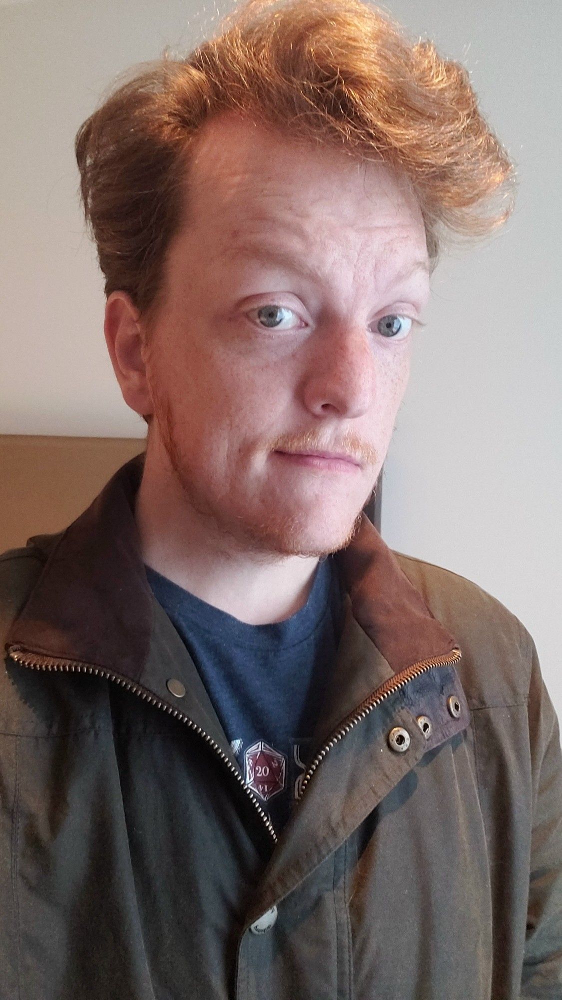 Selfie of a redheaded man wearing a navy shirt and an olive waxed cotton jacket. A red d20 design on the shirt is positioned at the vertex where the cost is zipped/buttoned.