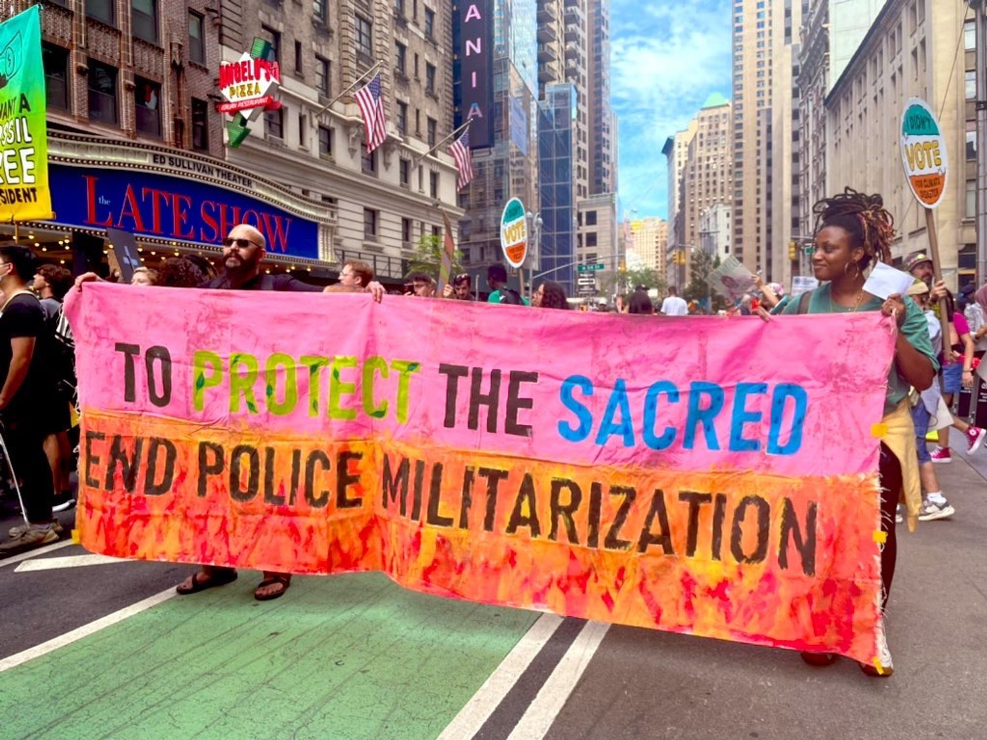 Pink banner reads “To Protect the Sacred END POLICE MILITARIZATION” with images of fire along the bottom edge. In the background you can see The Late Show studio.