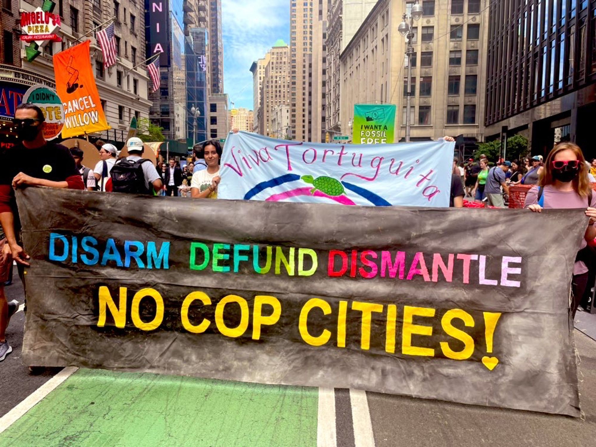 Two banners. One in rainbow letters on black background reads “Disarm. Defund. Dismantle” and underneath in yellow letters reads “No Cop Cities!” Second banner in the background on sky blue background, with green letters reads, “Viva, Viva, Tortuguita!” There is also a picture of a turtle. Thousands can be seen in the background on Broadway Ave.  with their own signs.