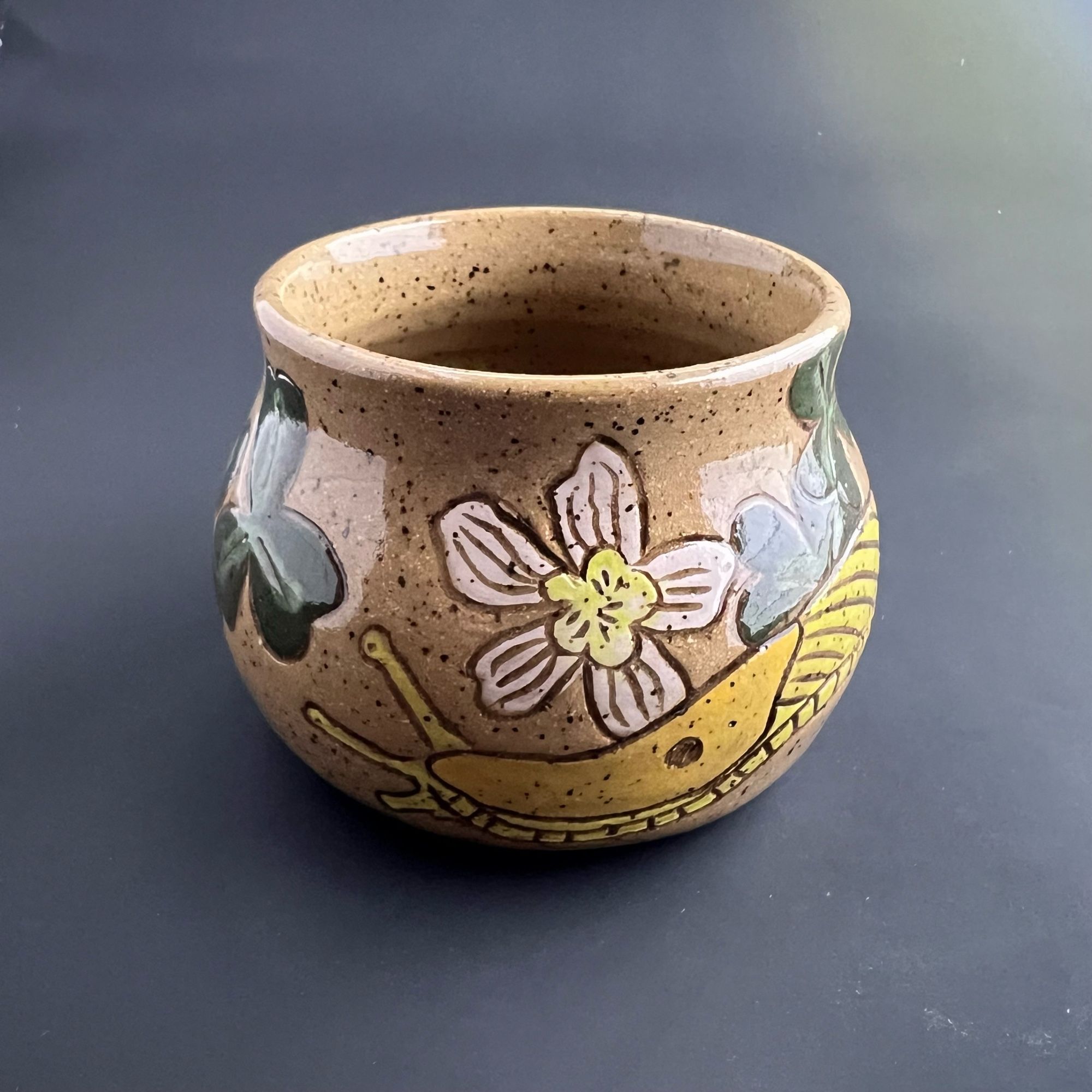 Small squat brown ceramic vessel with a banana slug design, accentuated by white and yellow flowers and clover-shaped leaves