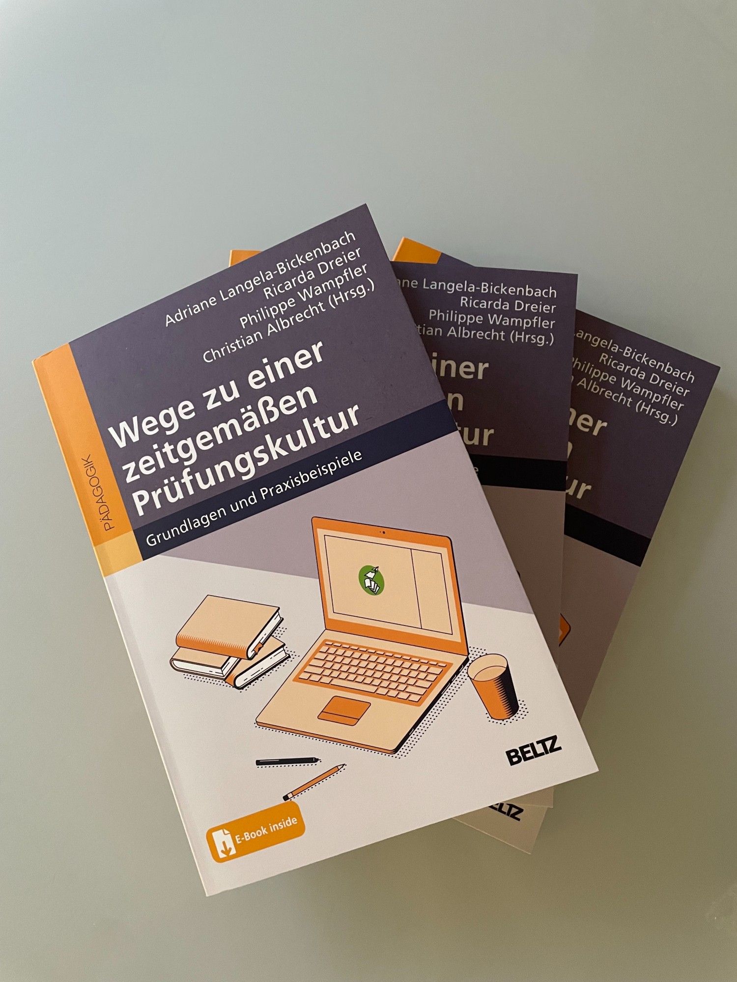 Das Bild zeigt drei Exemplare des Buches 'Wege zu einer zeitgemäßen Prüfungskultur' von Adriane Langela-Bickenbach, Ricarda Dreier, Philippe Wampfler und Christian Albrecht (Herausgeber:innen). Das Cover des Buches ist in blau und orange gehalten und zeigt neben einem Stapel Bücher noch ein aufgeklapptes Laptop, einen Becher mit einer hellen Flüssigkeit und zwei Stifte. Der Bildschirm des Laptops zeigt das Logo des Instituts für zeitgemäße Prüfungskultur.