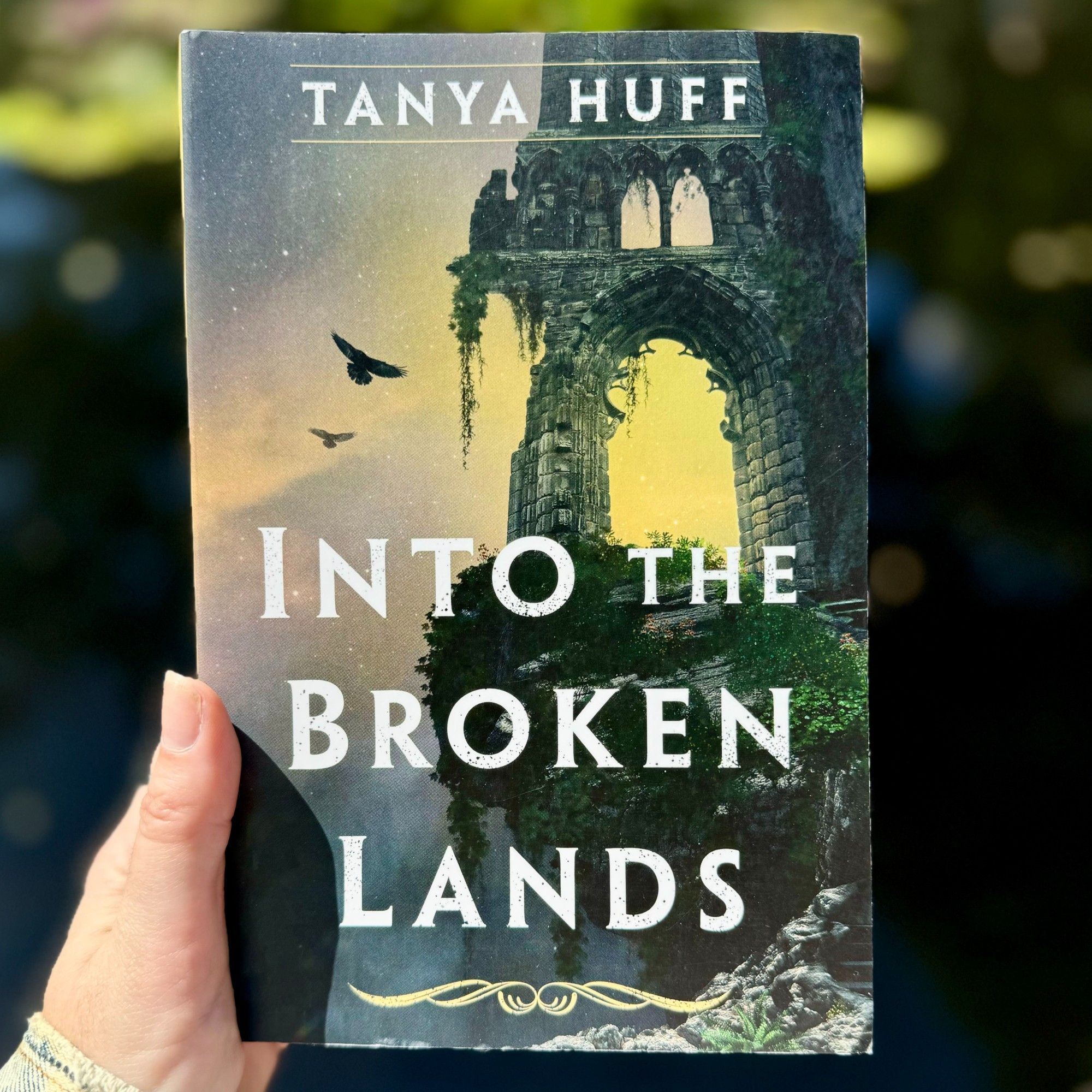 A hand holds up a hardcover copy of INTO THE BROKEN LANDS by Tanya Huff before a dark and shadowed greenery.