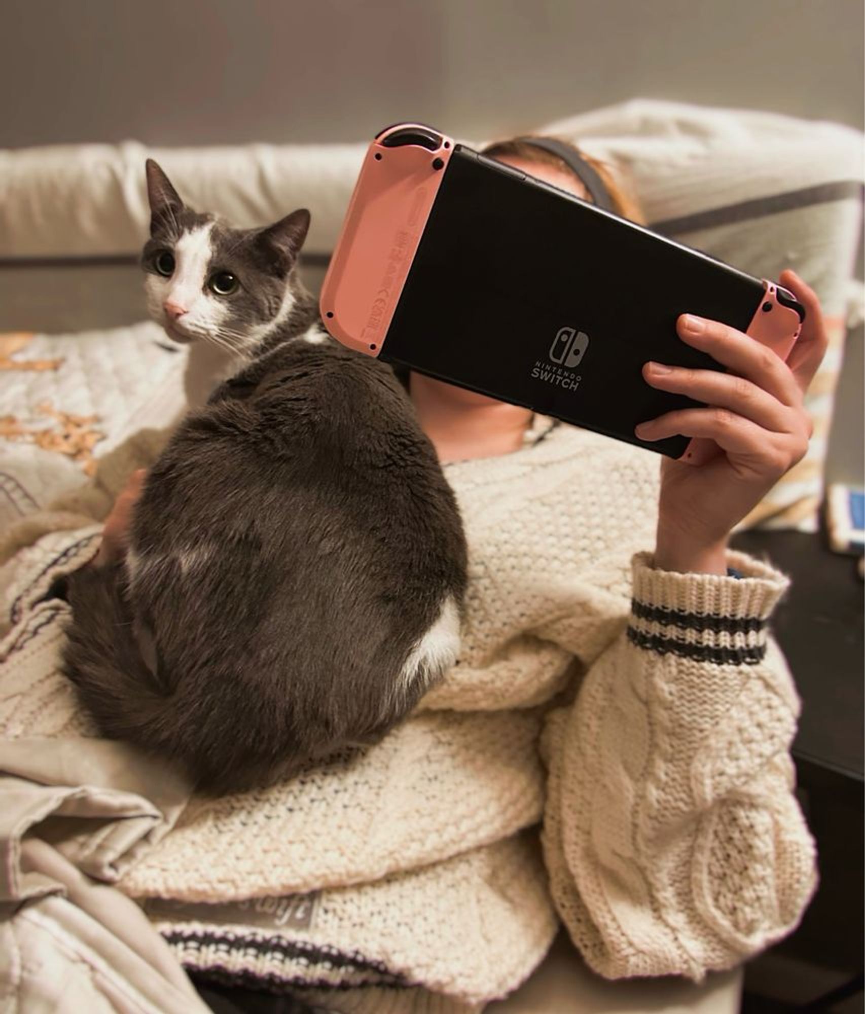 Photo of me laying in bed with my gray and white cat laying on my chest. My Nintendo switch is in my hand and is obscuring my face. 