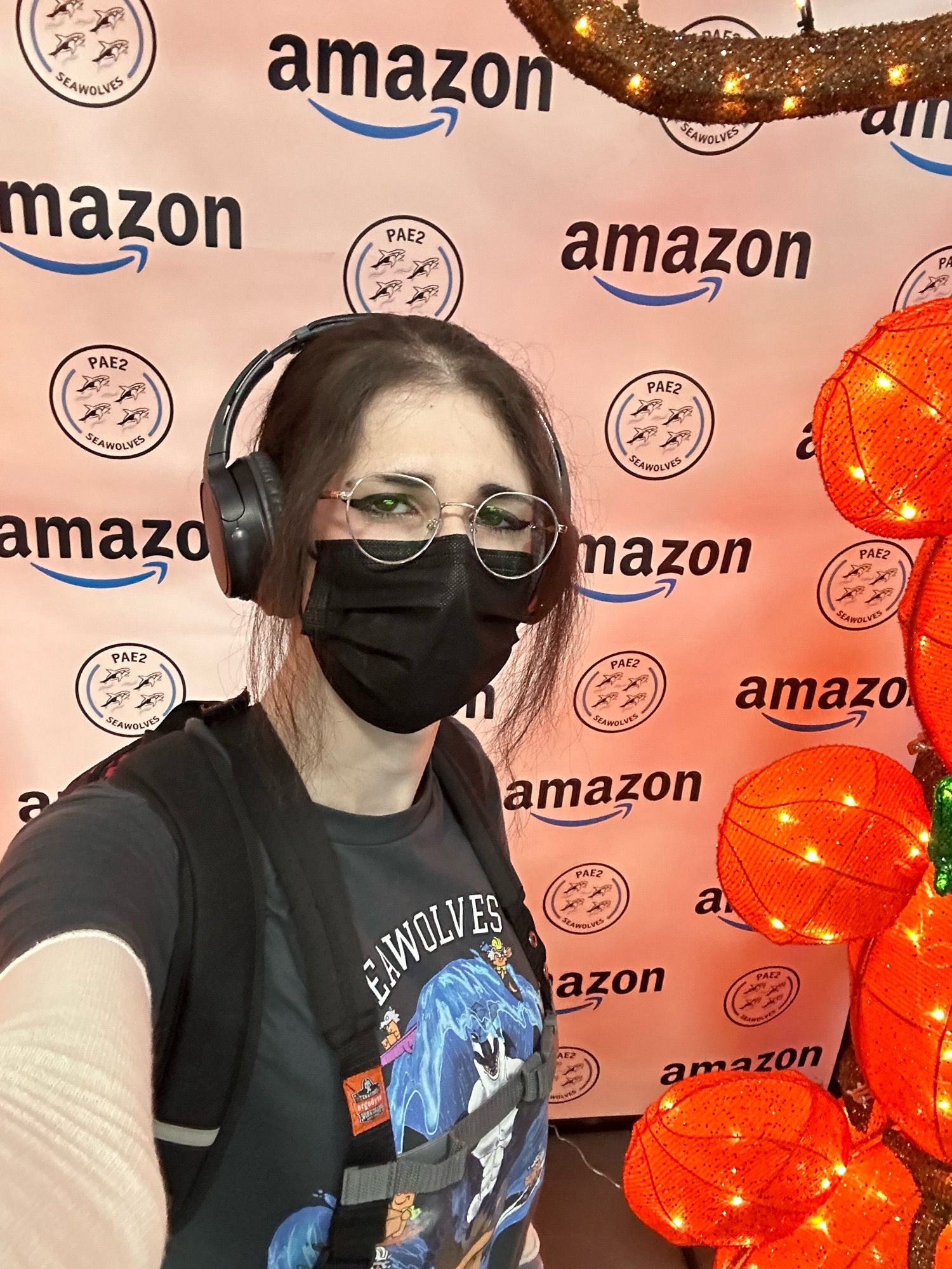 Beautiful woman with black hair tied up in that fuckass haircut Chell from portal has posing for a selfie in front of an arc of fake glowing pumpkins with an Amazon logo backdrop. Also she has big dumb round glasses and did I mention very pretty and cute also her son is dead