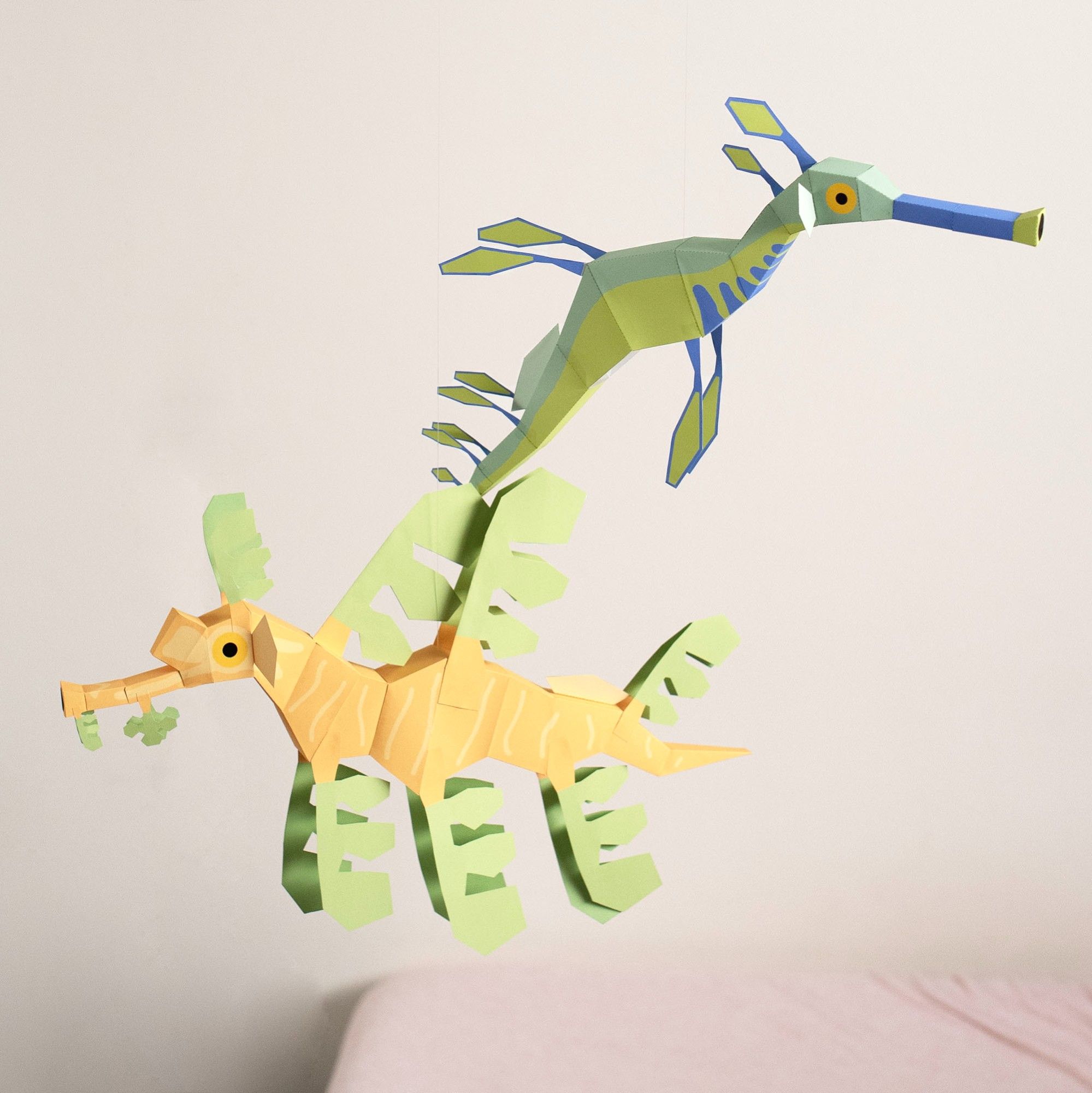 Two paper models hang suspended in the air against a white wall. On the upper right is a Weedy seadragon model, on the lower left is a Leafy seadragon model.