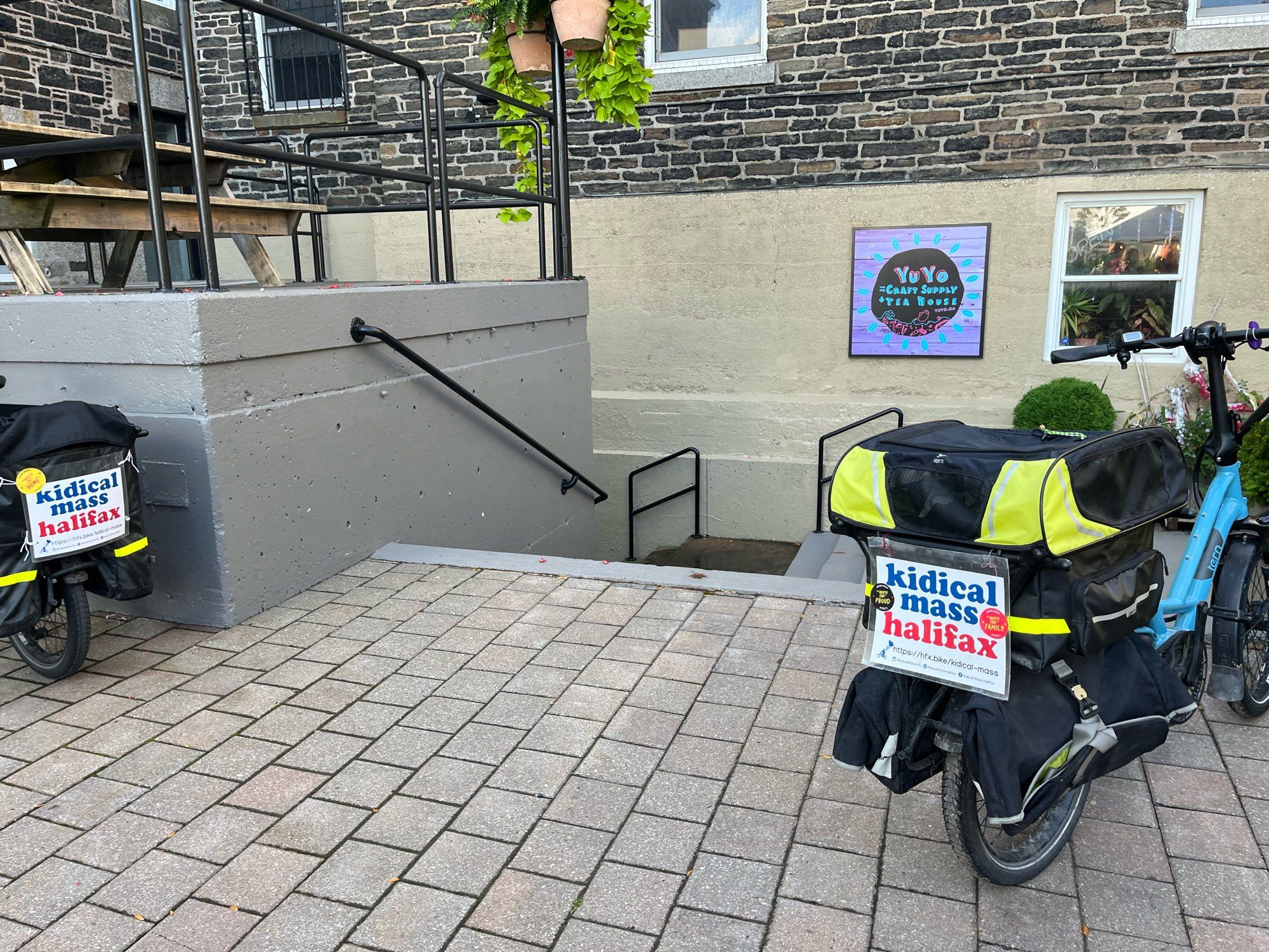 Two electric cargo bikes parked on a brick plaza. The back of both bikes have signs on them advertising Kidical Mass Halifax (and added stickers on them for Stephen MacKay running for election in District 8).