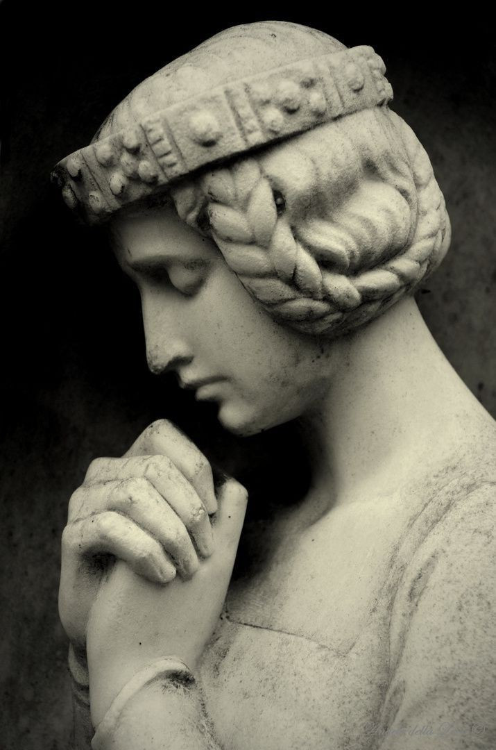 A monochromatic photograph of a statue of a woman, with braided hair and a headband-like crown, praying silently with her eyes closed and hands in front of her chest