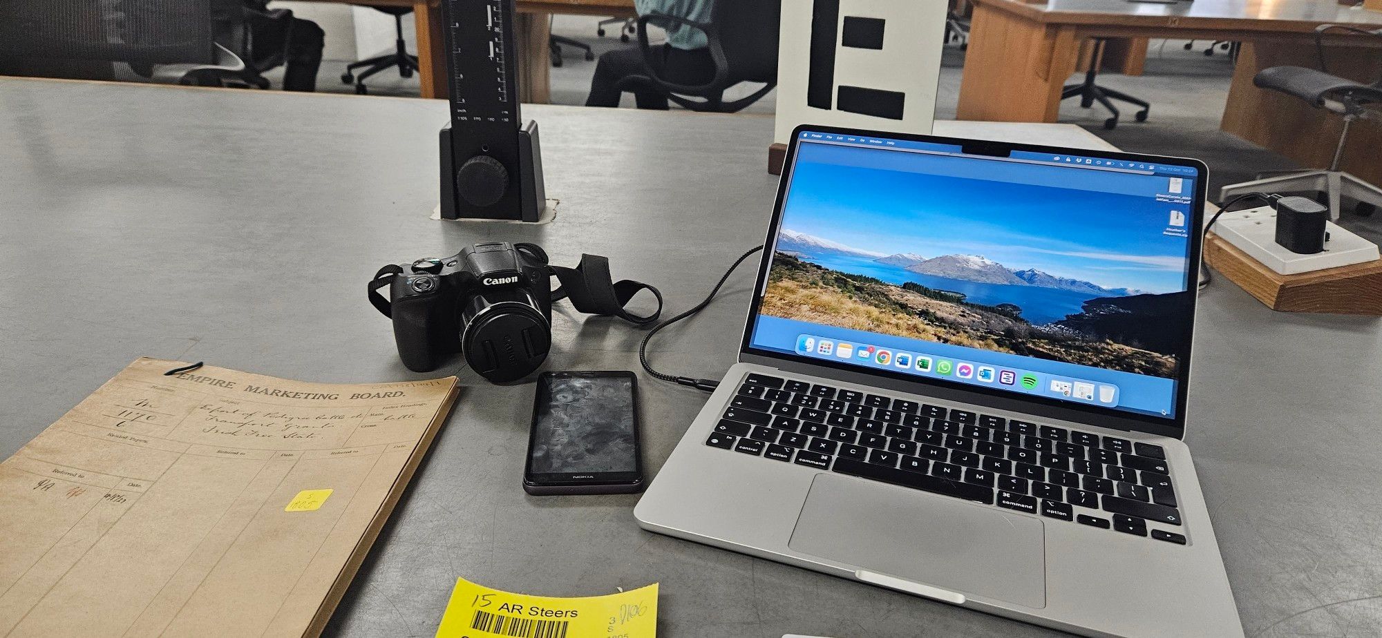 A brow n folder document, a Canon camera, phone, readers ticket and MacBook on a desk