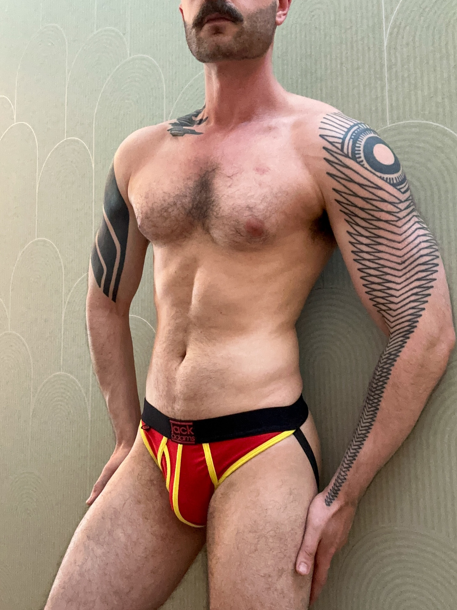 Me wearing a red and yellow jockstrap in front of a green pattered wall. 