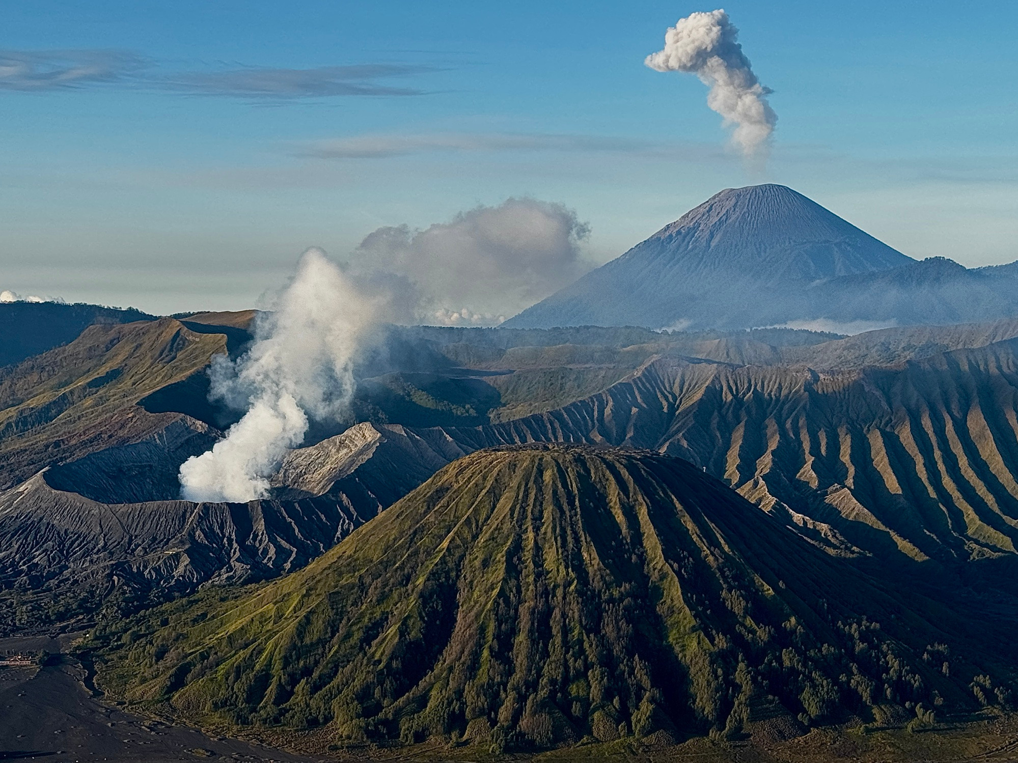 In East Java, Indonesia, volcanic ridges carpeted in green rise and fall like waves across the frame, as twin volcanoes exhale in unison — Bromo low and smouldering, Semeru high and commanding.