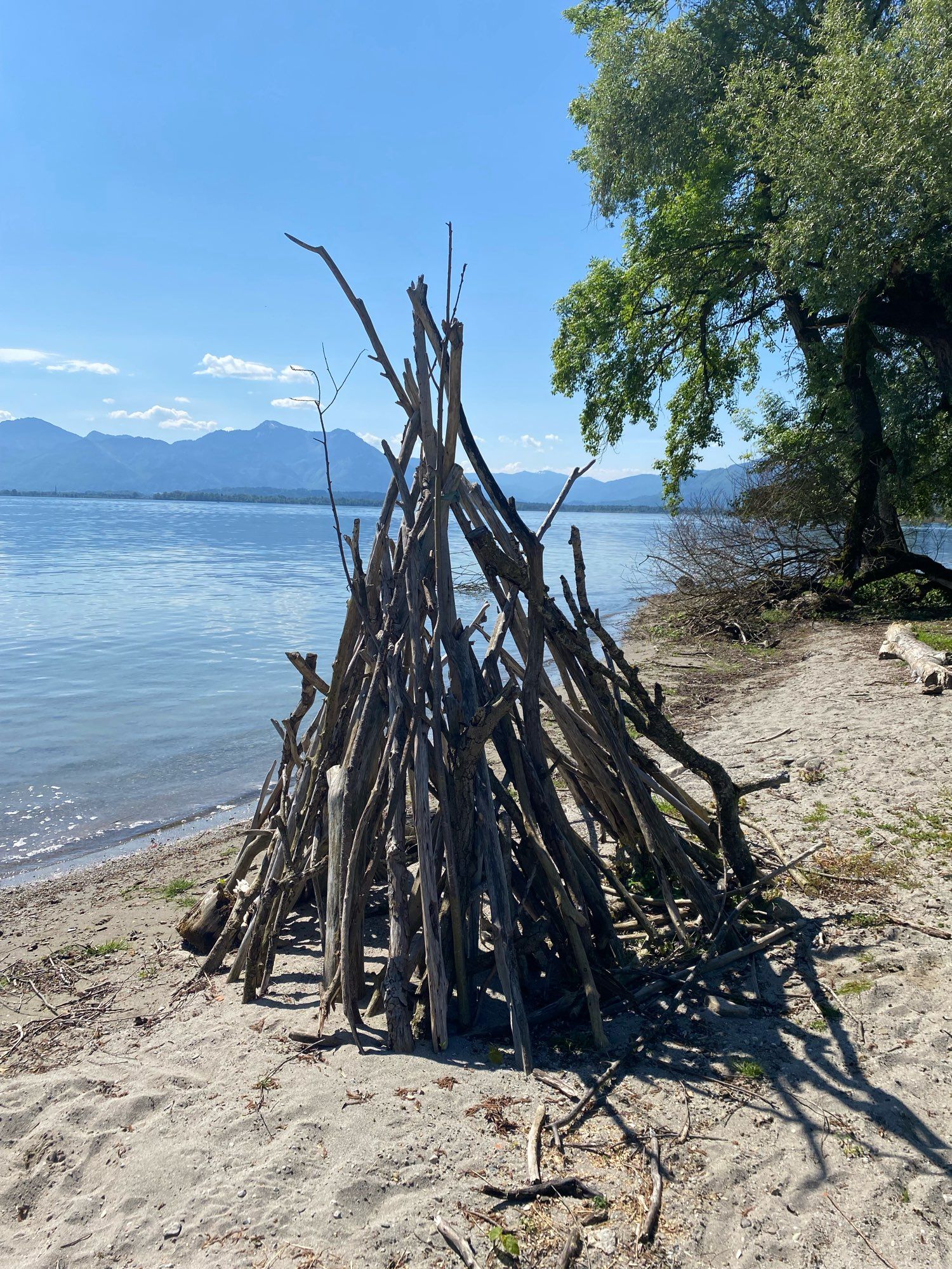 Sandiges Ufer mit Tipi aus Treibholz. See und Bergkulisse im Hintergrund.