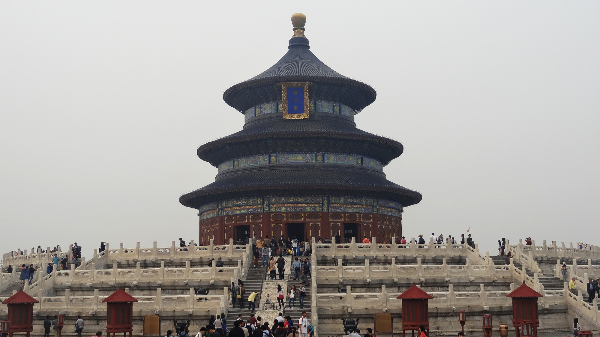 La photo montre le Temple du Ciel à Pékin, en Chine. C'est un complexe architectural circulaire à plusieurs niveaux, dominé par des couleurs vives. Des milliers de visiteurs montent les marches en marbre menant à l'édifice, entouré de balustrades ornées. Le ciel gris ajoute une atmosphère mystérieuse à la scène.