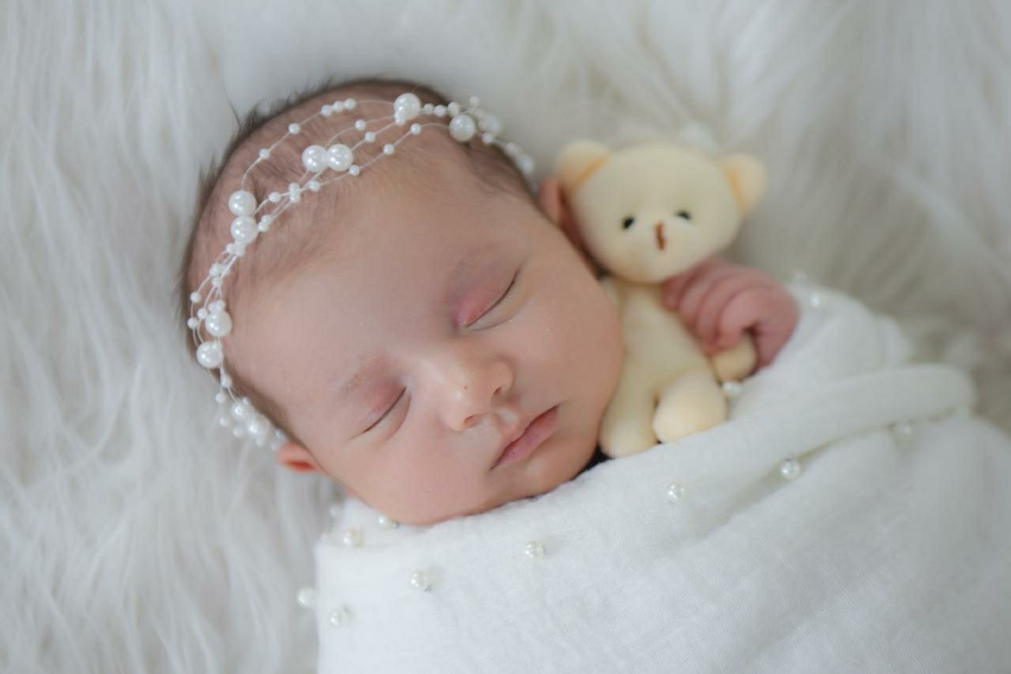 uma linda neném recém nascida enrolada num paninho branco bordado com pérolas, segurando um ursinho pequeno. a neném linda está de olhinhos fechado e também usa uma tiara de pérolas.