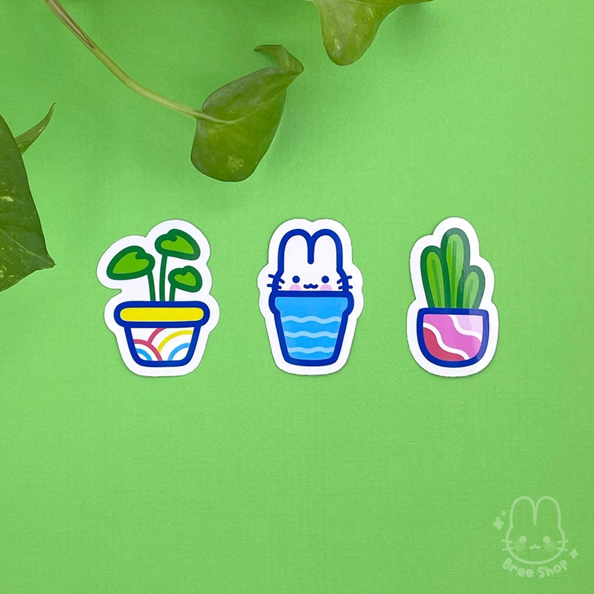 Photo of three stickers laying on a green background. Each sticker is a different illustrated colorful potted plant, the one in the middle has a smiling white bunny popping up out of it instead of a plant.