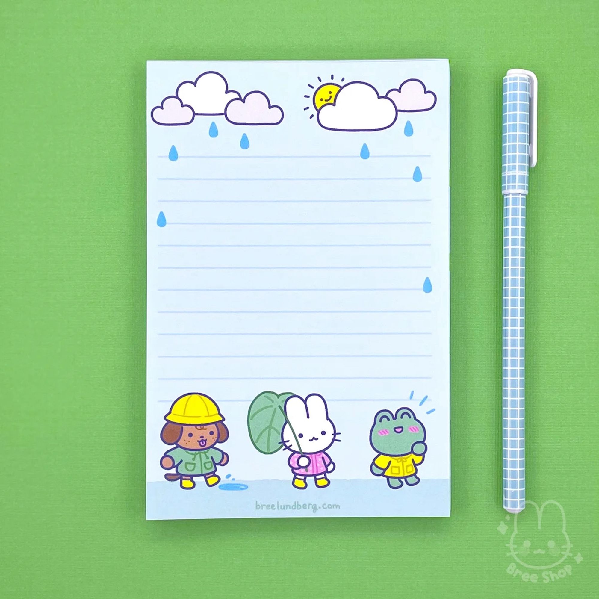 Photo of a light blue lined notepad on a green background. The notepad design has three cute characters dressed in rain gear (a puppy, bunny, and frog) along the bottom and clouds and raindrops along the top.