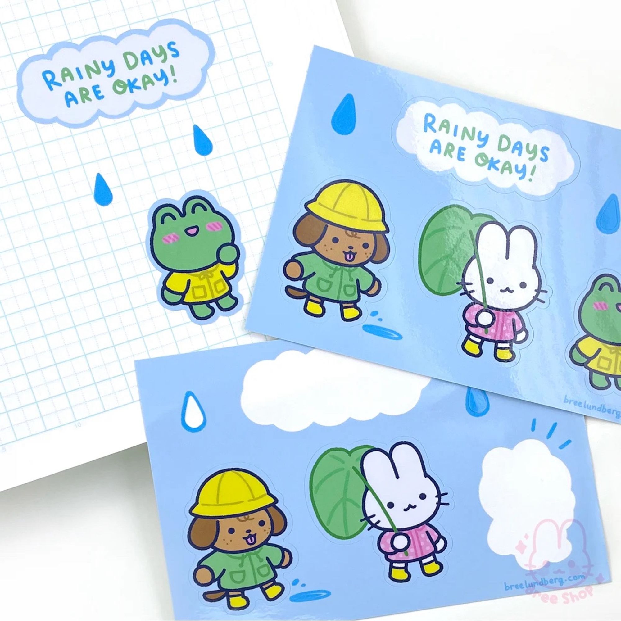 Photo of light blue sticker sheets with three cute characters dressed in rain gear (a puppy, bunny, and frog) along with raindrops and a cloud that says “rainy days are okay!”.