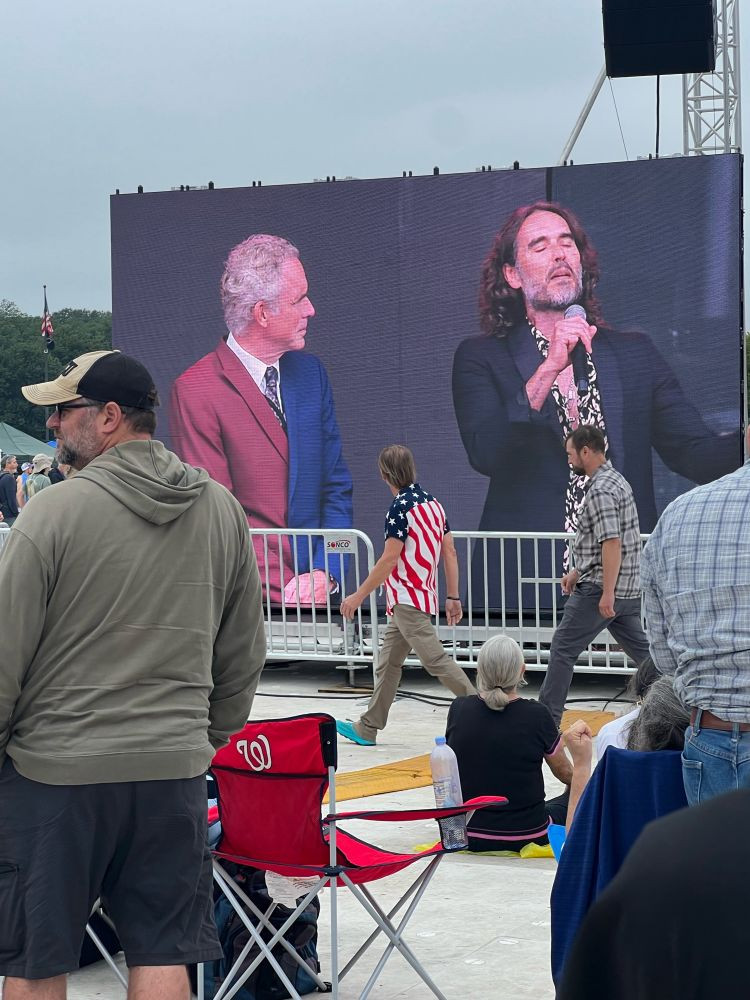 a screen outdoors showing Jordan Peterson looking at Russell Brand