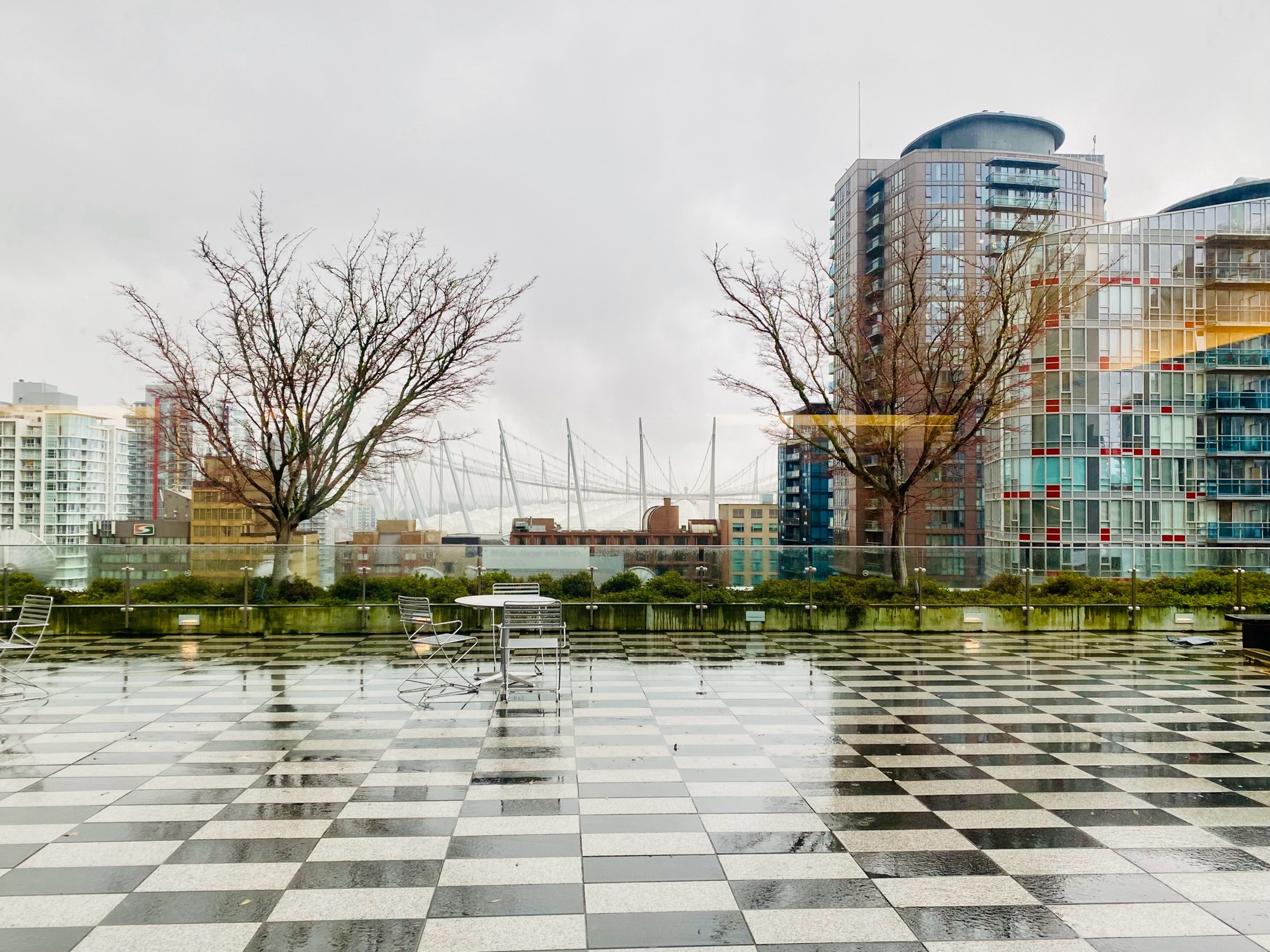 View from the Central Branch of the Vancouver Public Library on a rainy day. Chequered floor, a few trees, and the spikes of BC stadium in the background.