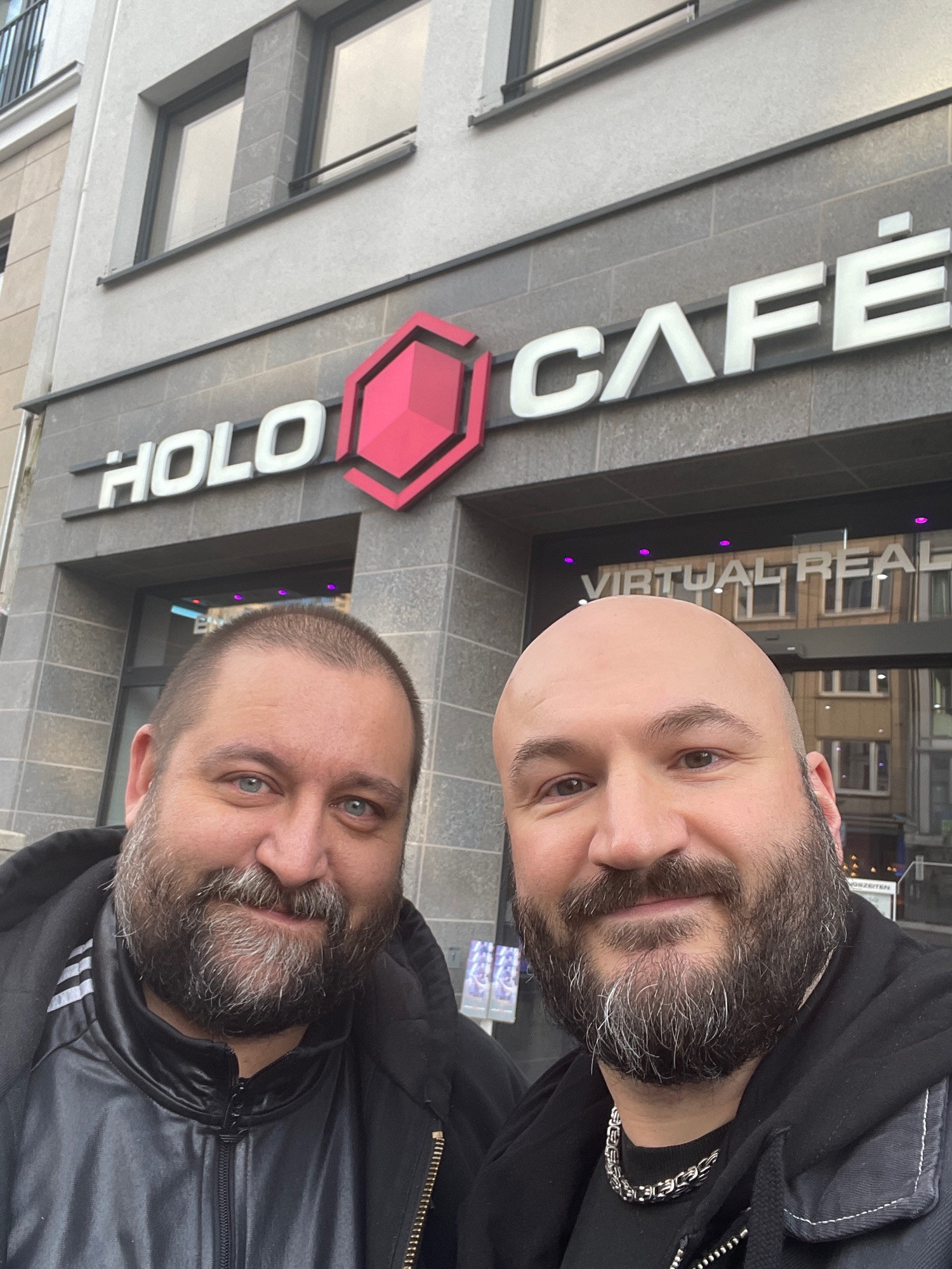 Selfie of two bearded guys in their early fourties. The left guy on the picture with a buzz cut the right guy bald and both in front of the words ”Holo Café“.