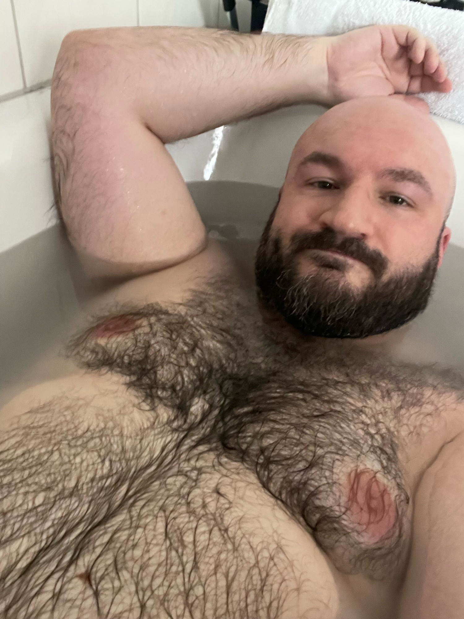 A 40 year old, bearded man in a bath tub from the belly upwards, showing hair on belly and chest. One arm is resting on his bald head.