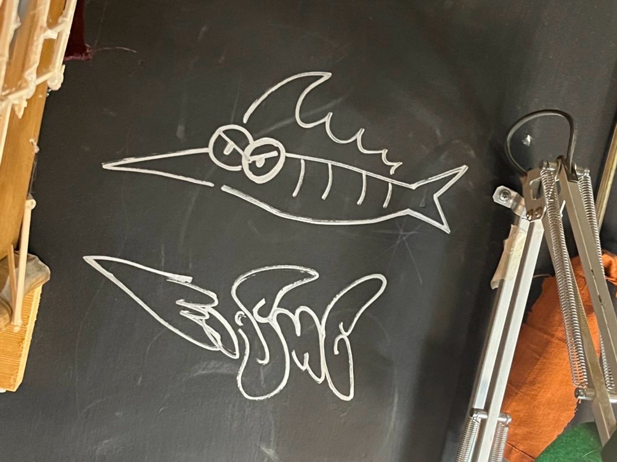 Graffiti on a dark wall, the upper part is a doodle is a simplistic marlin, the bottom part is a tag that reads “Fishe” that has a silhouette meant to resemble a marlin.