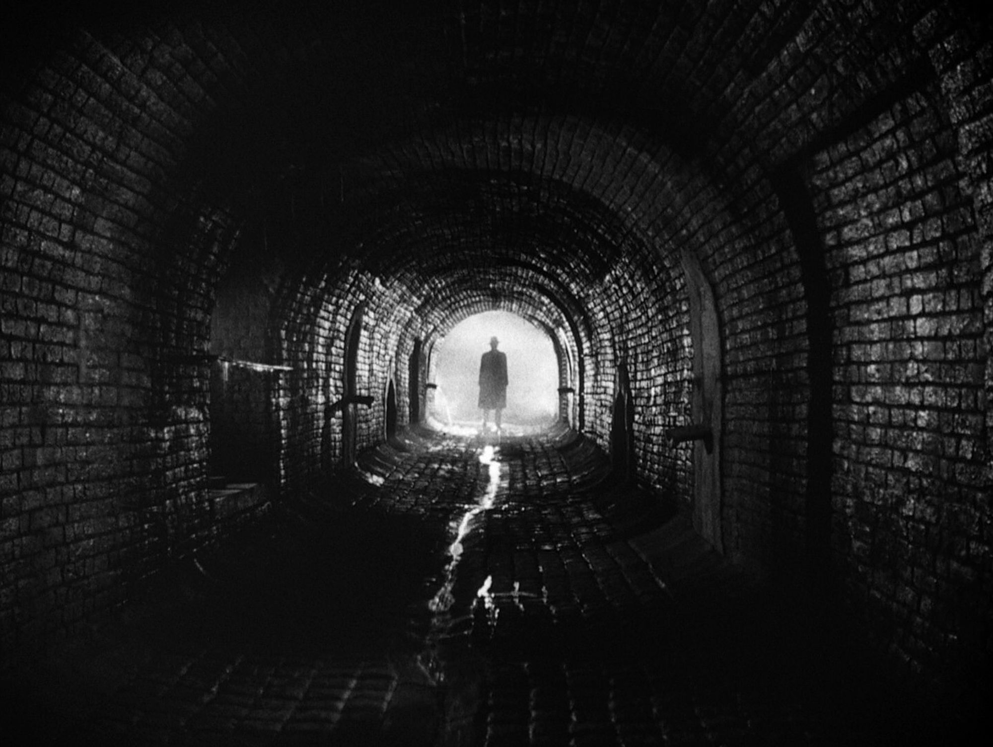 Black & white shot from the film "The third man", showing the silhouette of a man standing at the end of an underground tunnel.