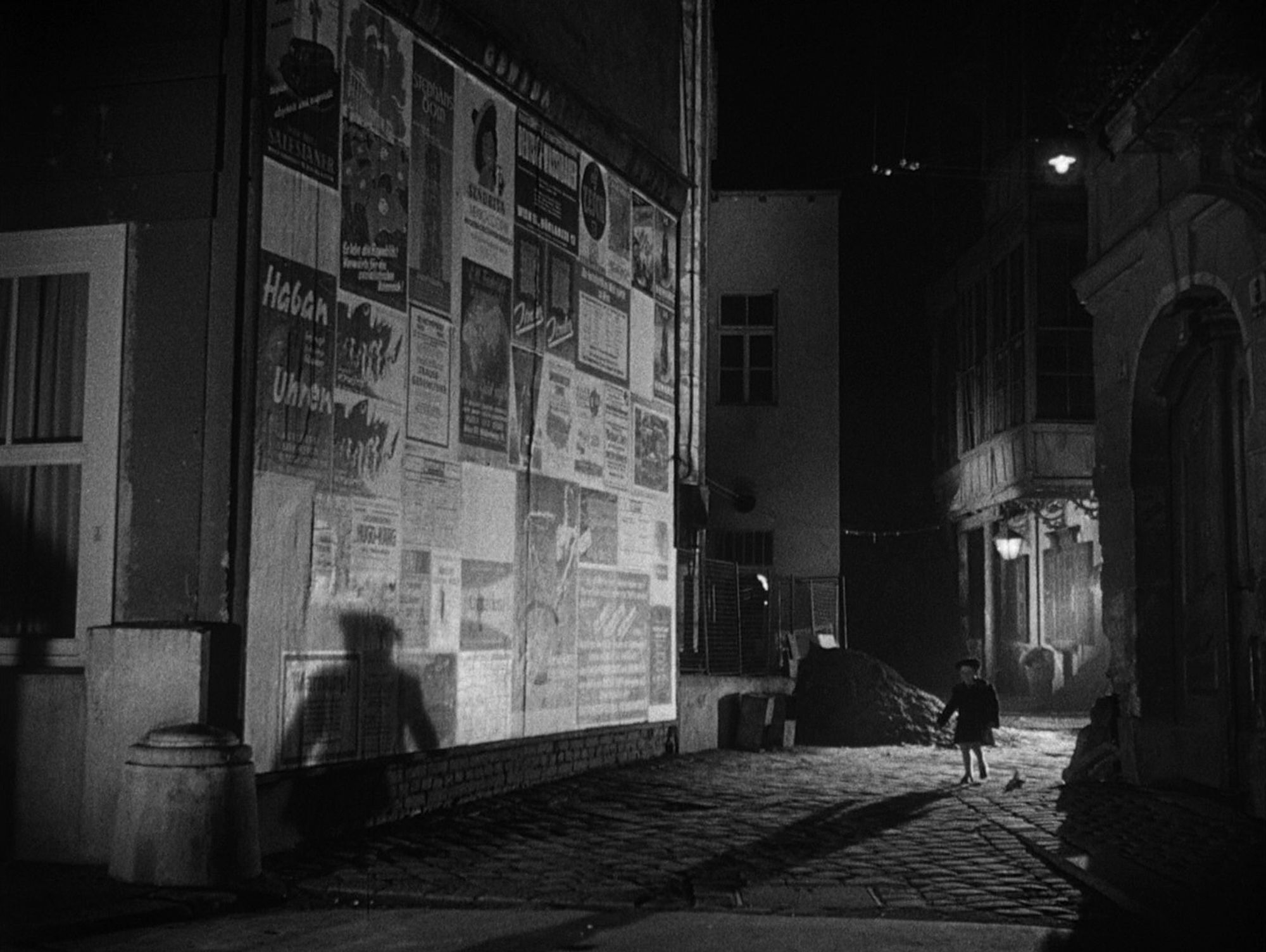 Black & white shot from the film "The third man", showing the silhouette of a child in a street in post-war Vienna. It is night and the child's shadow is projected onto a wall full of advertising posters.