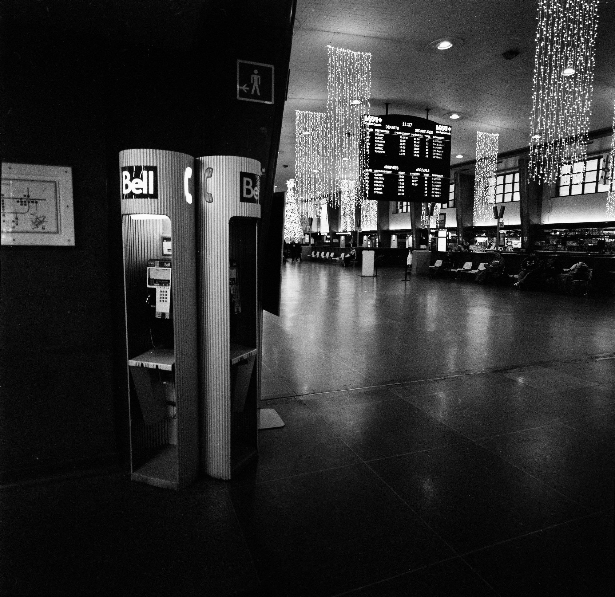 Two cylindrical payphone booths made of corrugated metal in a mostly empty train hall. The light in the one on the right is burned out. A large arrivals and departures board is in the distance.